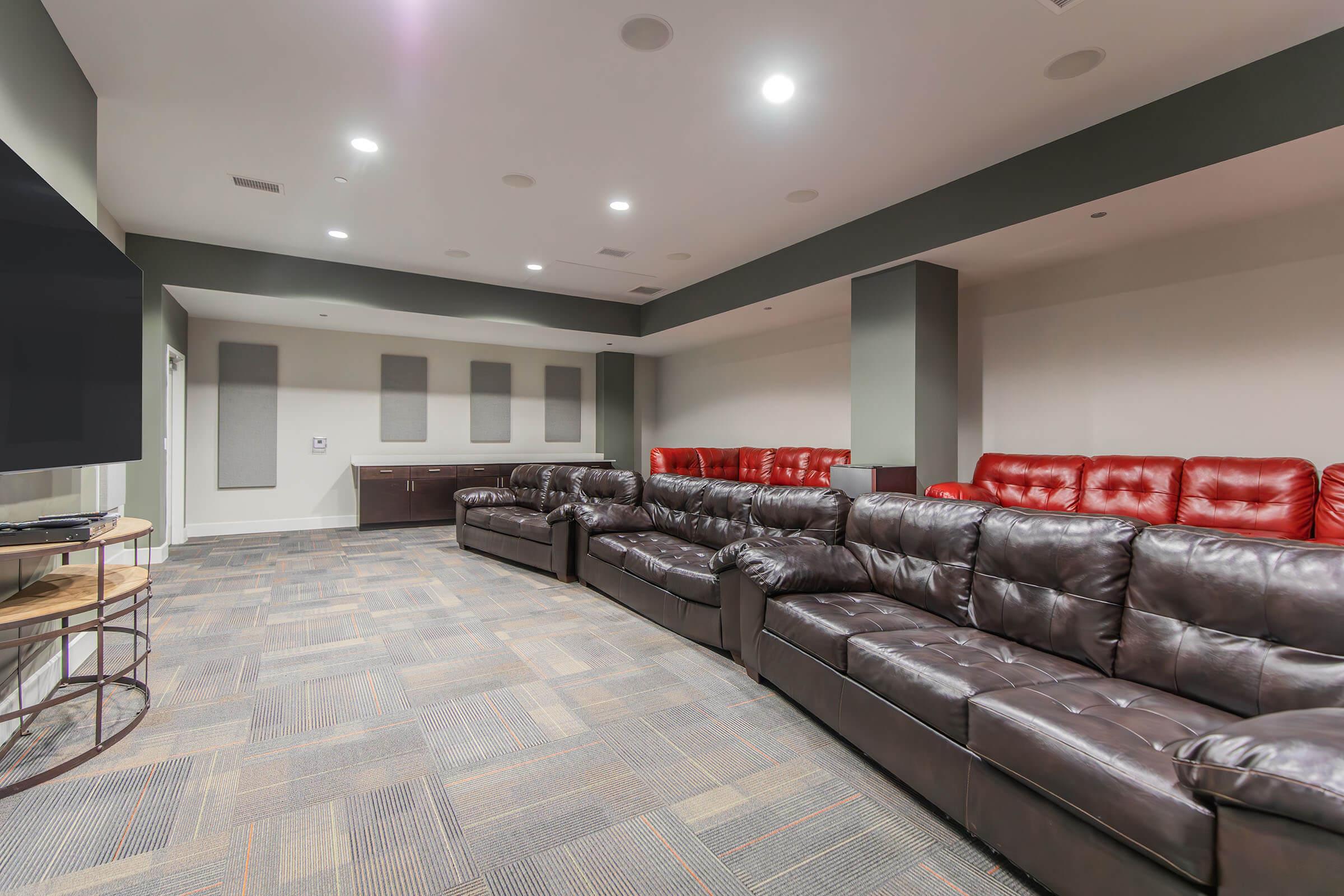 A spacious entertainment room featuring a large sectional sofa with dark brown and red leather seating. The room is well-lit with recessed lighting, has a large wall-mounted TV, and soundproof panels on the walls. The flooring is carpeted in a patterned design, creating a cozy atmosphere.