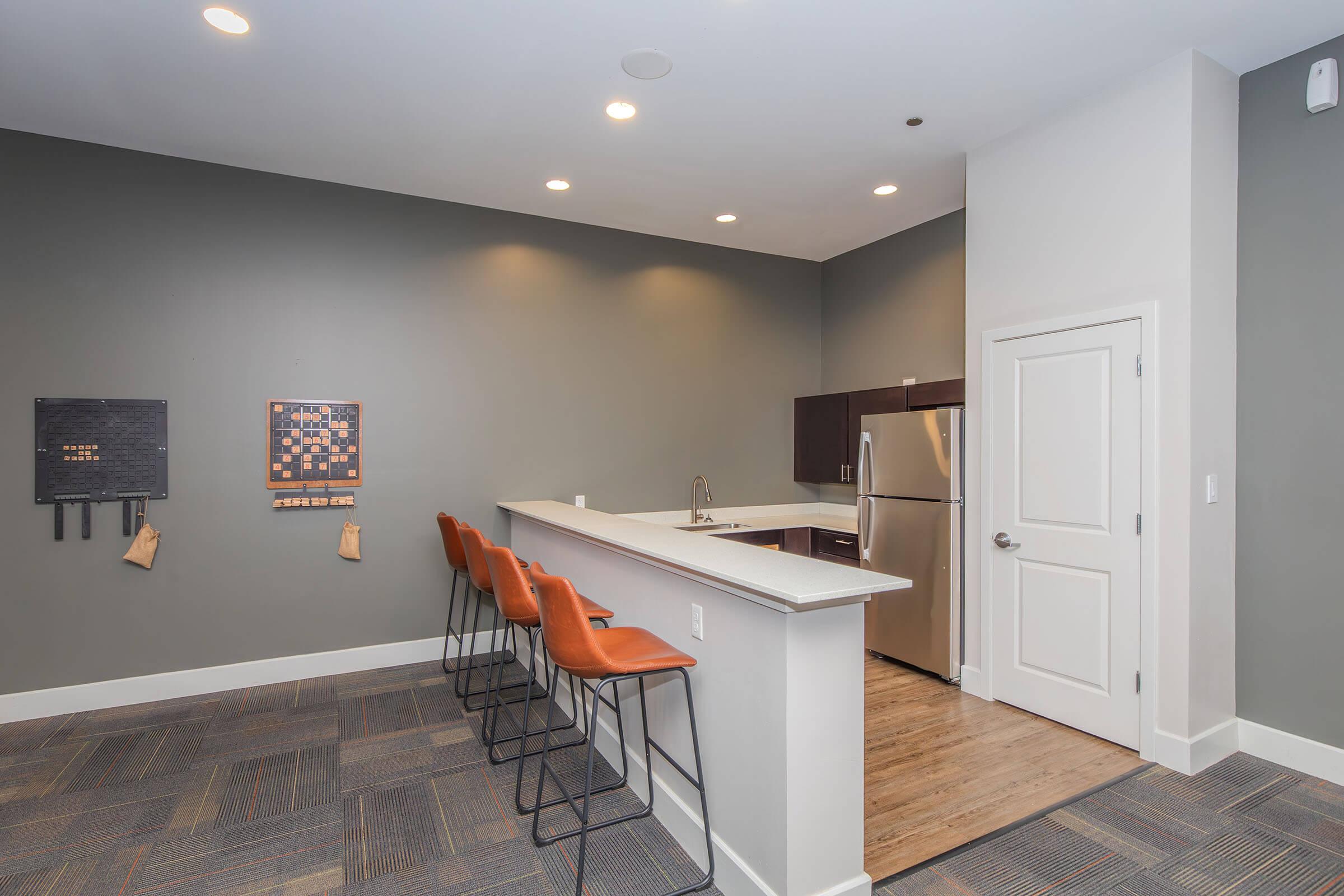 Modern kitchen and lounge area featuring a bar with four orange stools, a white countertop, and stainless steel appliances. The walls are painted grey, with a decorative wall art piece on one side and wood flooring. Bright LED lighting enhances the spacious, inviting atmosphere.