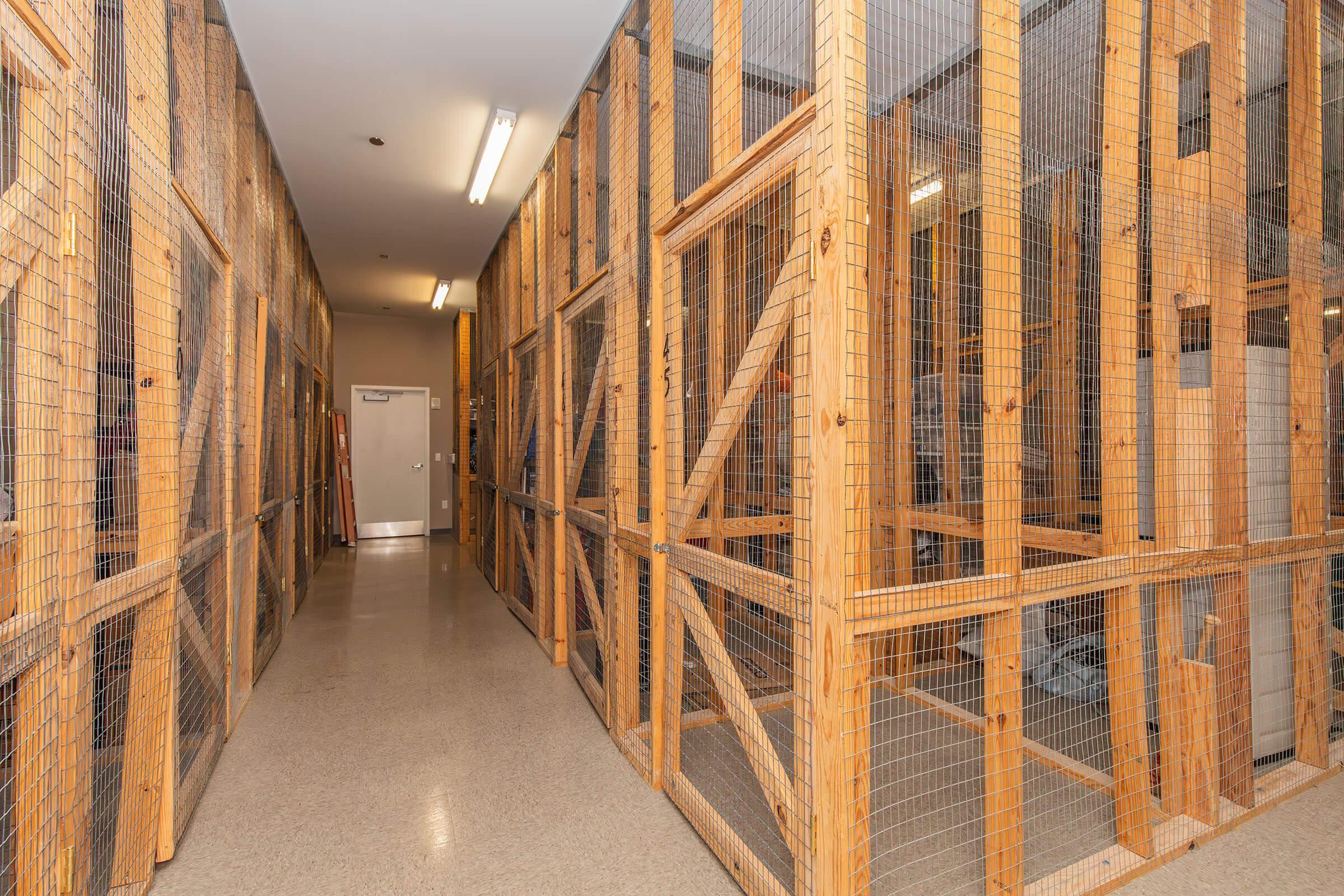 Interior view of a hallway lined with wooden cages or enclosures, featuring metal wire mesh and wooden frames. The space has overhead lighting and a door visible at the end of the corridor. The floor is smooth and appears clean, suggesting a facility designed for animal housing or storage.