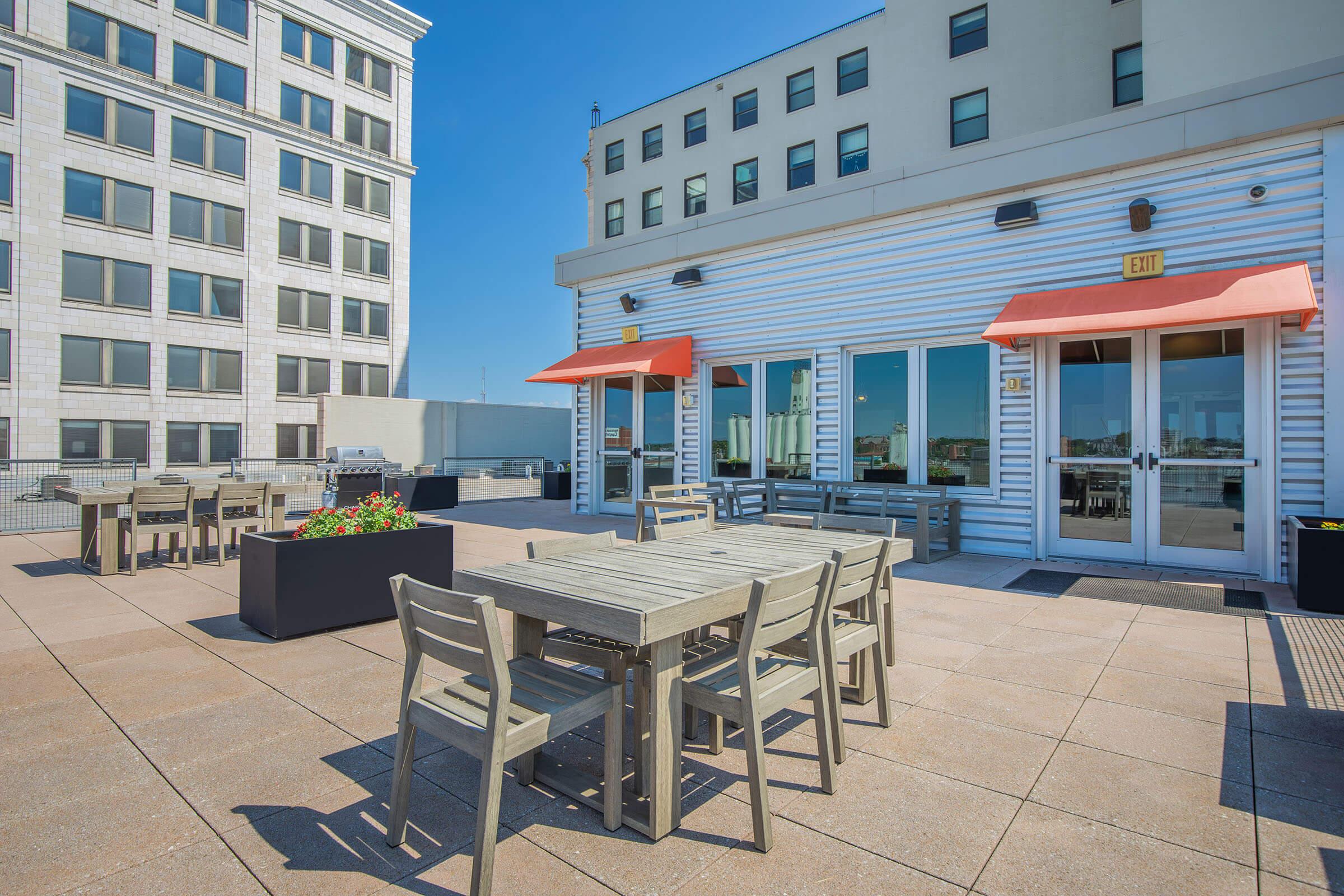 Rooftop terrace featuring multiple wooden tables and chairs, surrounded by flower planters. Two orange awnings provide shade over the seating area. In the background, a modern building with large windows and a clear blue sky can be seen.