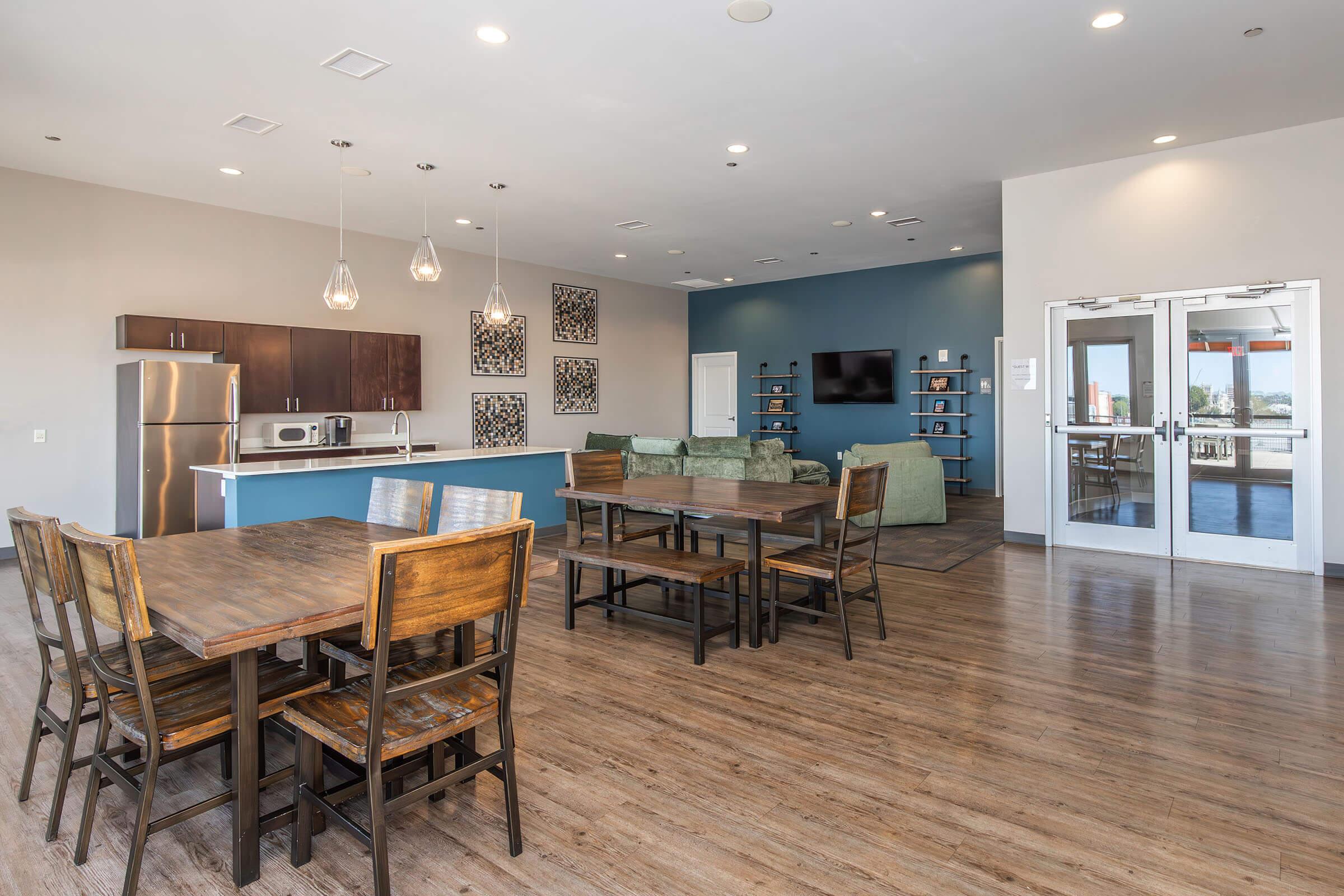Interior of a modern communal lounge area featuring a dining table with wooden chairs, a kitchen with dark cabinetry and stainless steel appliances, accented by decorative wall art. A soft seating area with a green couch and a television is visible, along with large windows allowing natural light.