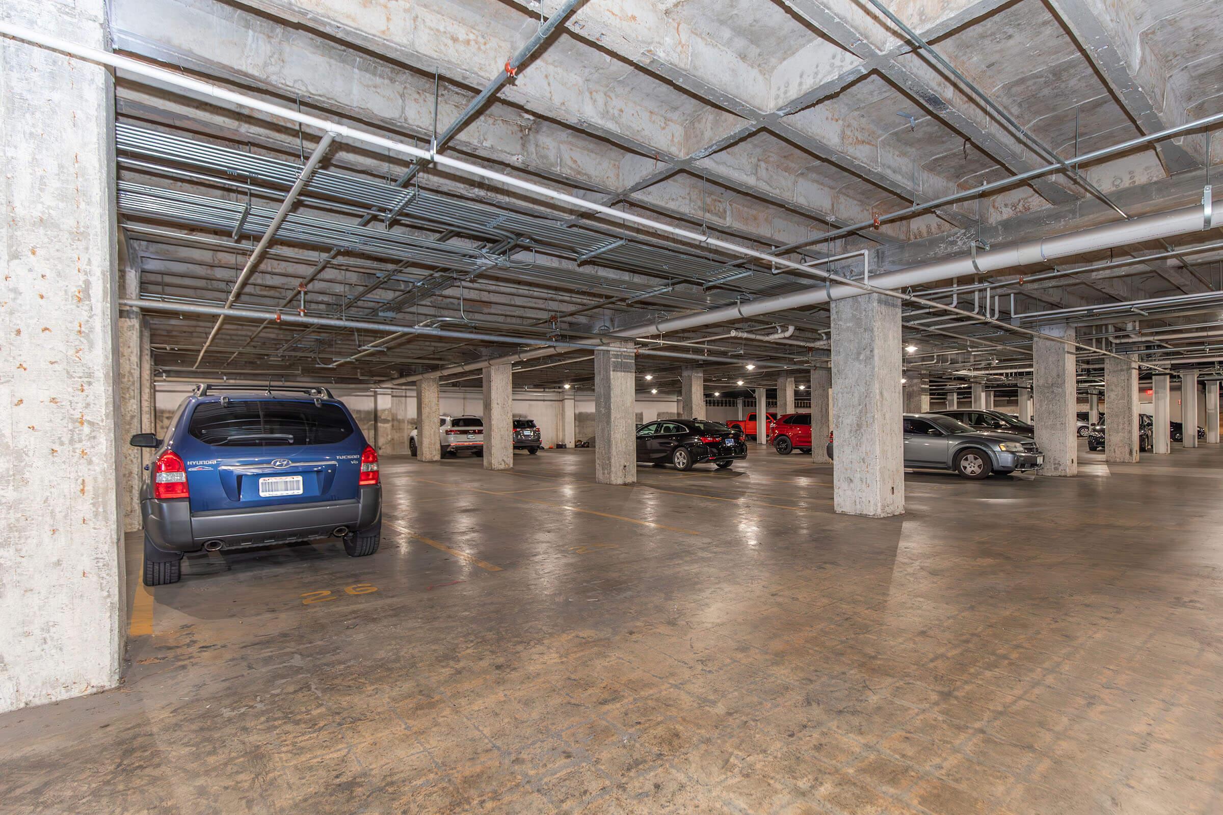A spacious underground parking garage with several parked cars, including a blue SUV, a black sedan, and a red vehicle. The concrete structure features exposed beams and pipes, with clear lines marking parking spaces. The floor is polished, and the lighting is bright, providing a well-maintained environment.