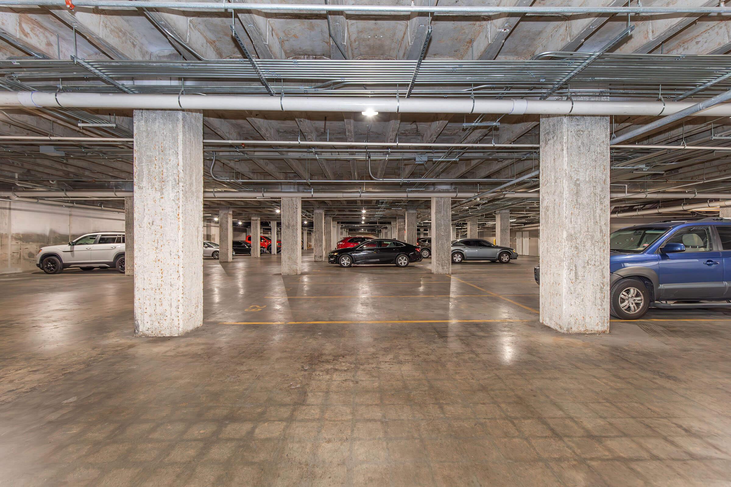 An empty underground parking garage with several parked vehicles. The space features concrete pillars, exposed overhead pipes, and a clean, well-lit environment. The floor has a grid pattern and there is a clear layout of parking spaces.