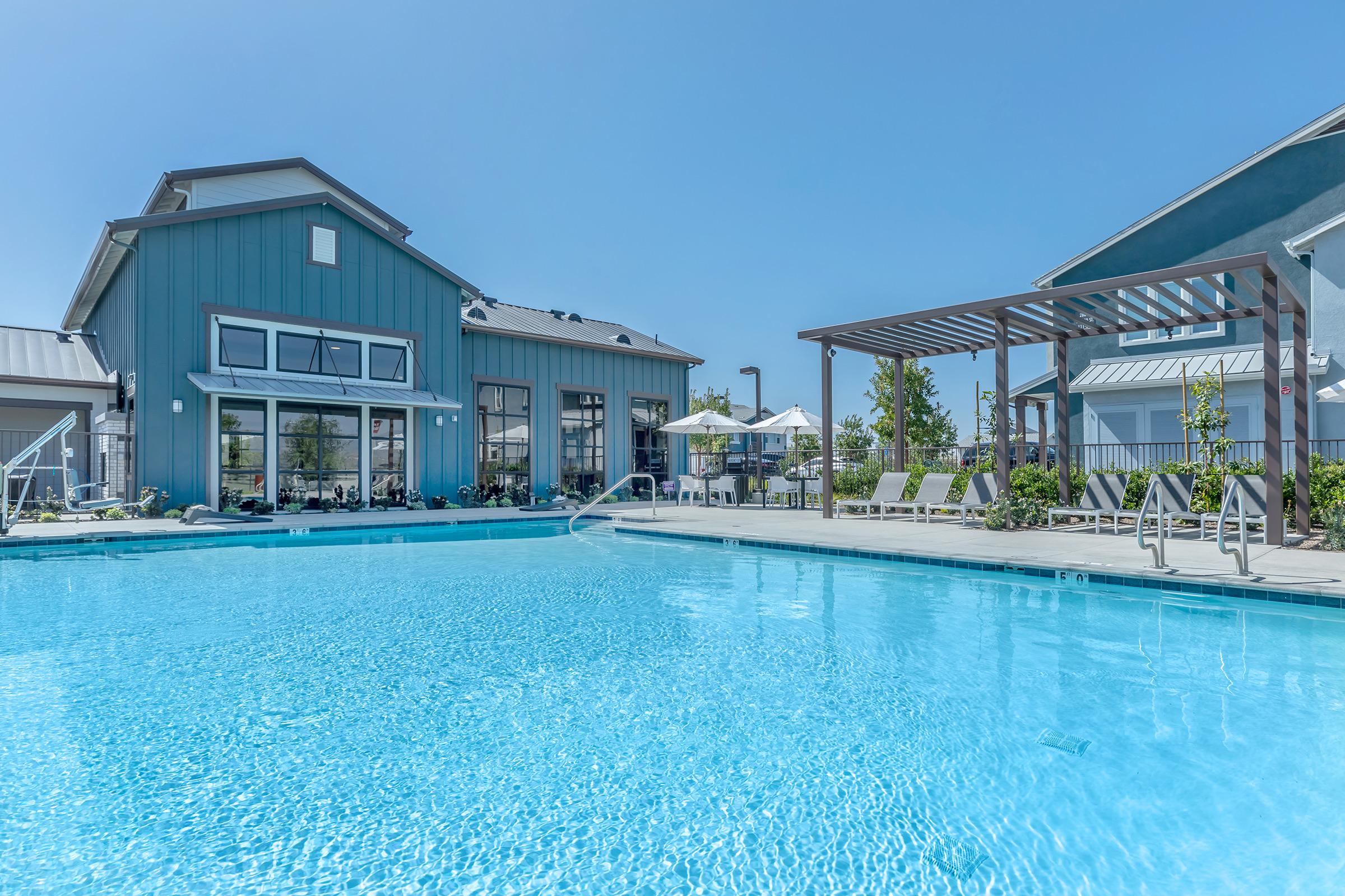 A bright blue swimming pool surrounded by modern buildings. Lounge chairs line the poolside, and a pergola with plants provides shade. Clear skies above create a sunny, inviting atmosphere, perfect for relaxation and leisure activities.