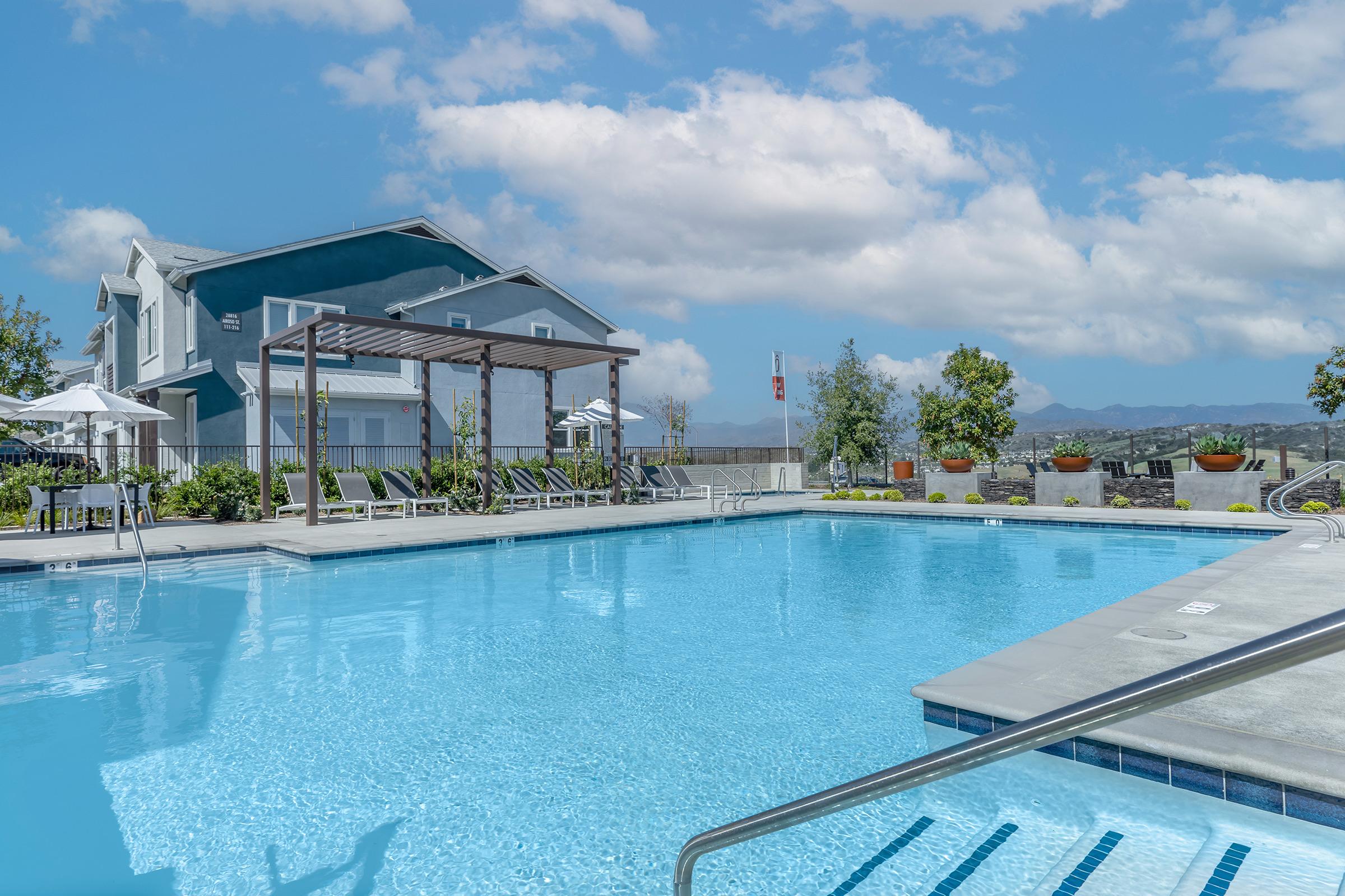 A clear blue swimming pool surrounded by lounge chairs and umbrellas, with a modern house in the background. Lush greenery and potted plants are placed around the pool area, and the sky is partly cloudy, creating a serene and inviting atmosphere.