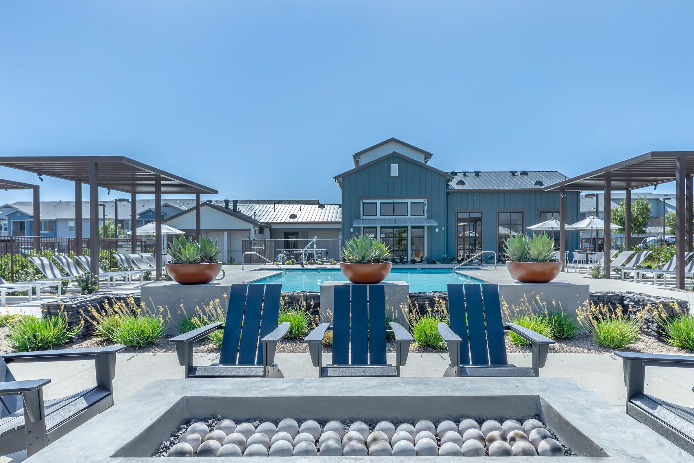 A serene outdoor pool area featuring lounge chairs and potted plants. In the background, there is a stylish building with a metal roof. Sun loungers are arranged around the pool, creating a relaxing atmosphere. Clear blue skies add to the inviting ambiance.