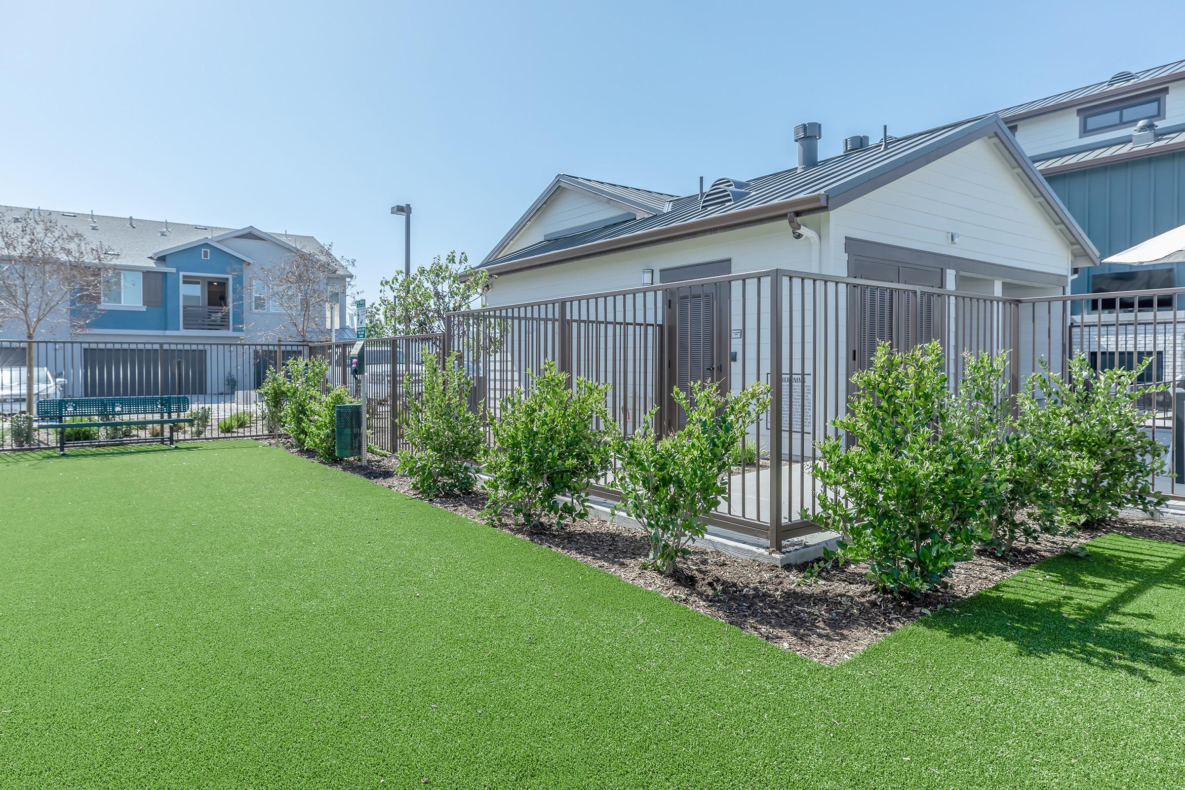 A well-maintained outdoor area featuring artificial turf, surrounded by low greenery. In the background, there are modern buildings, and a small fenced structure is visible. The scene is bright and sunny, creating an inviting atmosphere for leisure activities.