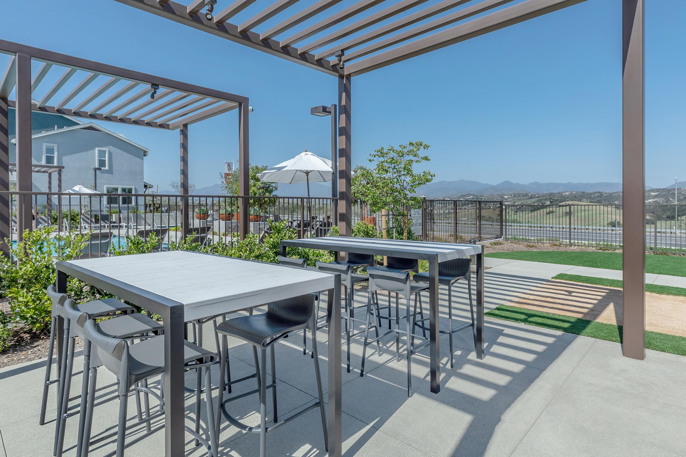 Outdoor patio area with modern tables and chairs under a pergola, overlooking a scenic landscape with mountains in the background. Features include a grassy area and an umbrella for shade, creating a relaxed setting ideal for gatherings or dining. Clear blue sky enhances the tranquil atmosphere.