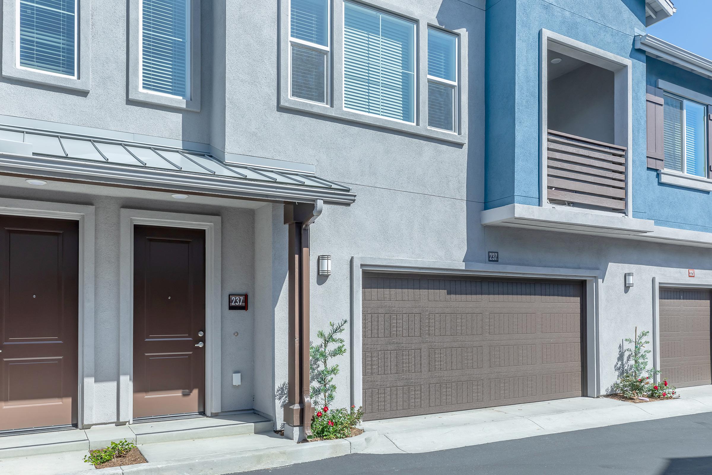 A modern multi-unit building with a gray exterior and blue accents. The image shows two entry doors, each with a number, and matching garage doors below. The walkway is lined with small plants, and there are windows above with horizontal blinds. Bright, sunny day with clear skies.