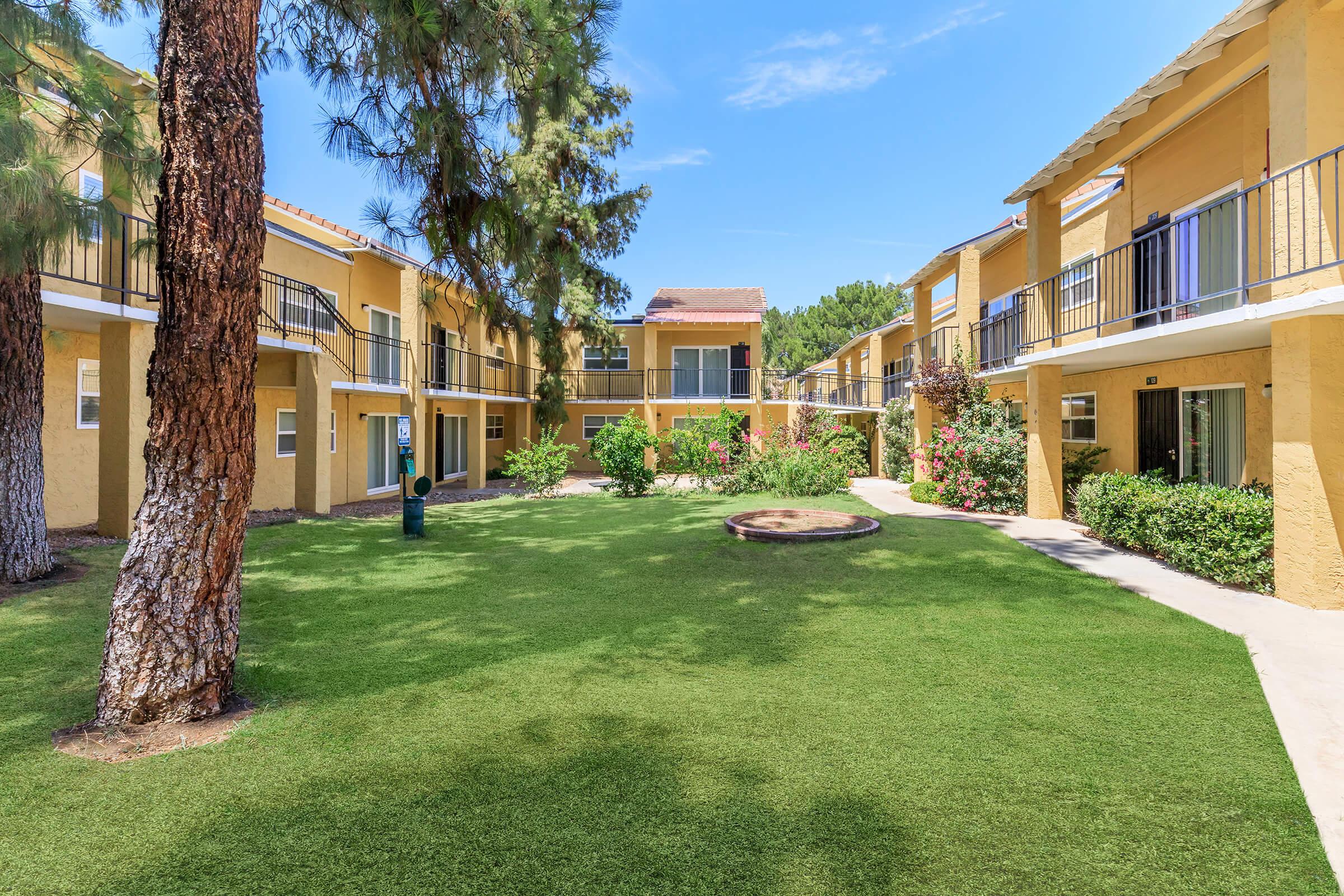 A well-maintained courtyard surrounded by yellow apartment buildings. Lush green grass covers the ground, and colorful flowerbeds add natural beauty. Tall trees provide shade, and a circular seating area is visible in the center, creating a welcoming outdoor space for residents.