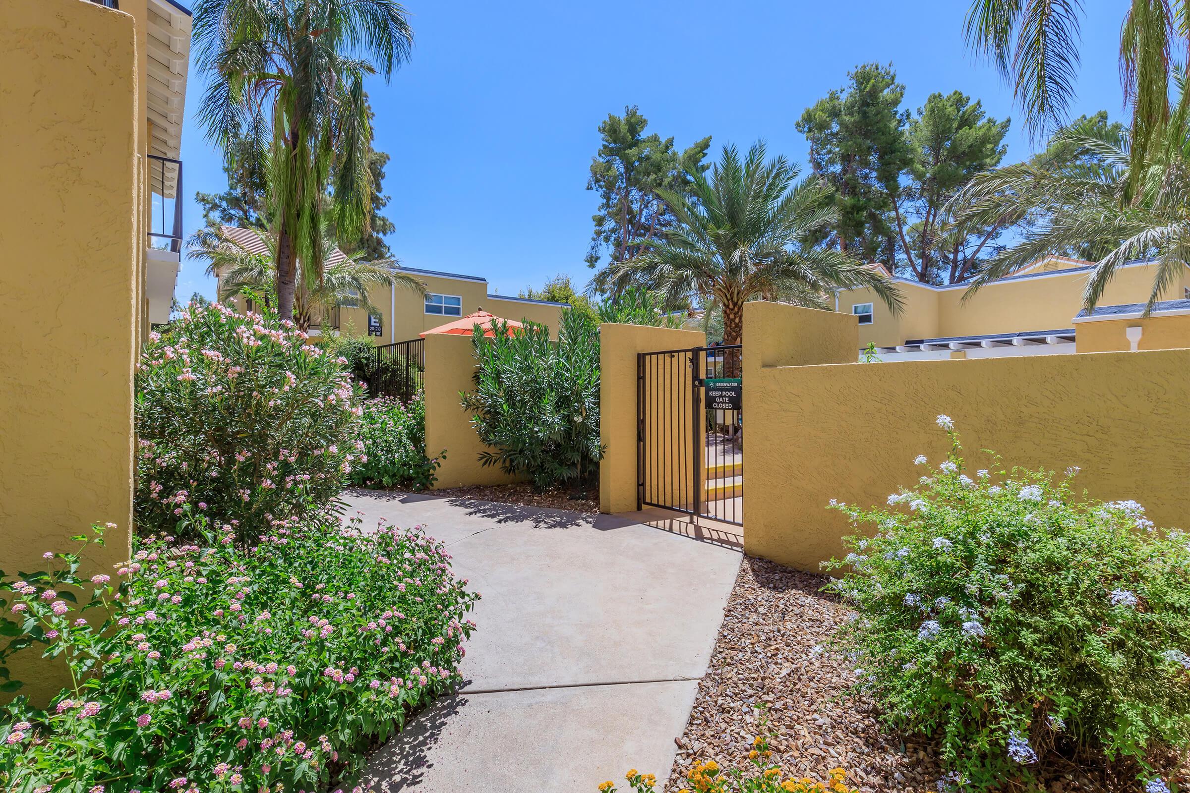 A sunny outdoor pathway leading through a garden filled with lush greenery and colorful flowers. On either side, there are tall palm trees and a gated entrance that opens to a pool area beyond the walls. The clear blue sky enhances the vibrant atmosphere of the setting.