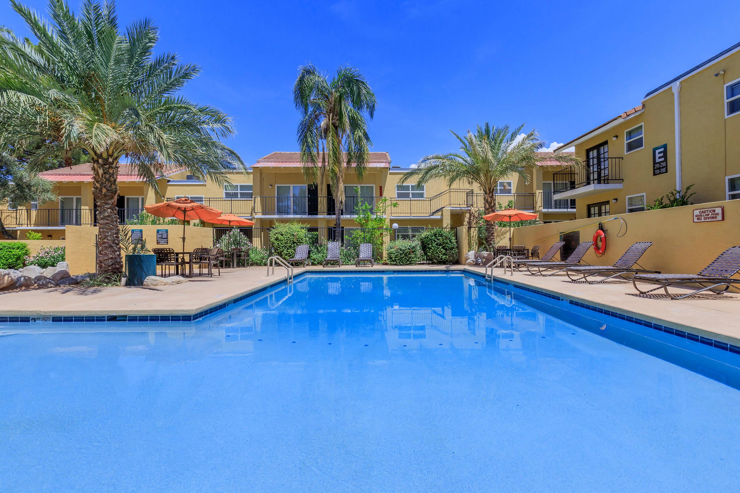 A sunny outdoor pool scene surrounded by palm trees and lounge chairs. The pool is clear blue and reflects the bright sky. In the background, there are buildings with balconies and outdoor seating areas under orange umbrellas. The area has a relaxed, tropical feel.