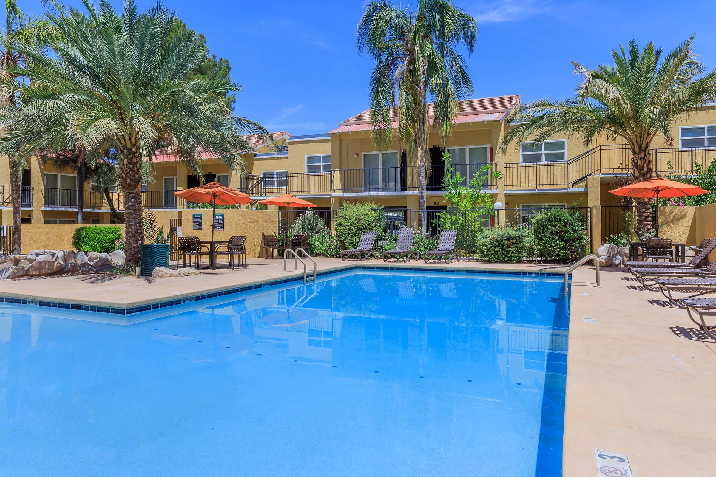 A serene pool area surrounded by palm trees and lounge chairs, featuring orange umbrellas for shade. The backdrop includes a multi-story building with balconies and greenery, all under a clear blue sky. Perfect for relaxation and leisure.