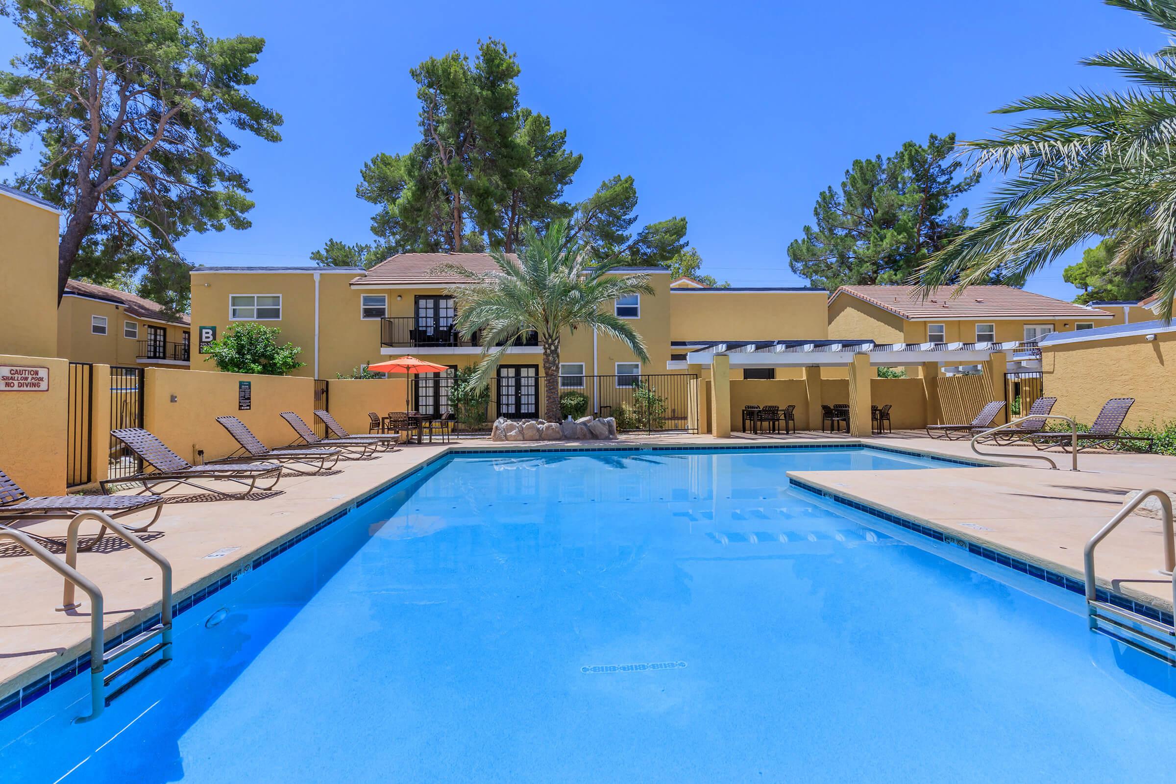 A clear blue swimming pool surrounded by lounge chairs, palm trees, and several buildings with yellow exteriors. The scene is sunny and inviting, featuring a shaded area with umbrellas and seating adjacent to the pool. Green foliage adds a tropical feel to the setting.