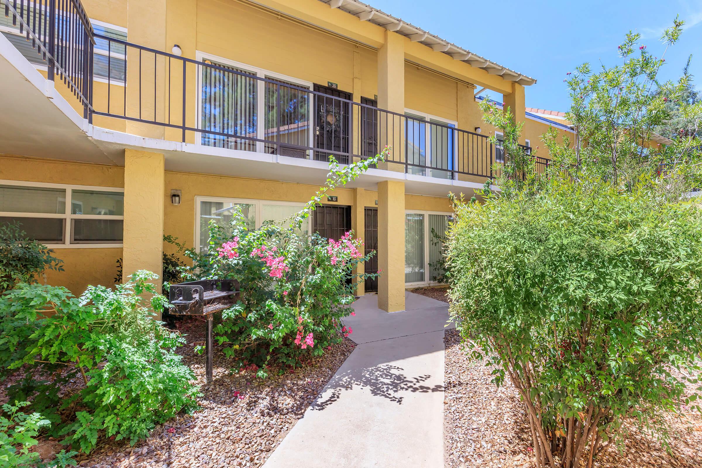 A yellow two-story apartment building with balconies, surrounded by lush greenery and flowering plants. A winding pathway leads to the entrance, with a barbecue grill visible in the garden area. Bright blue sky above adds to the cheerful ambiance of the scene.