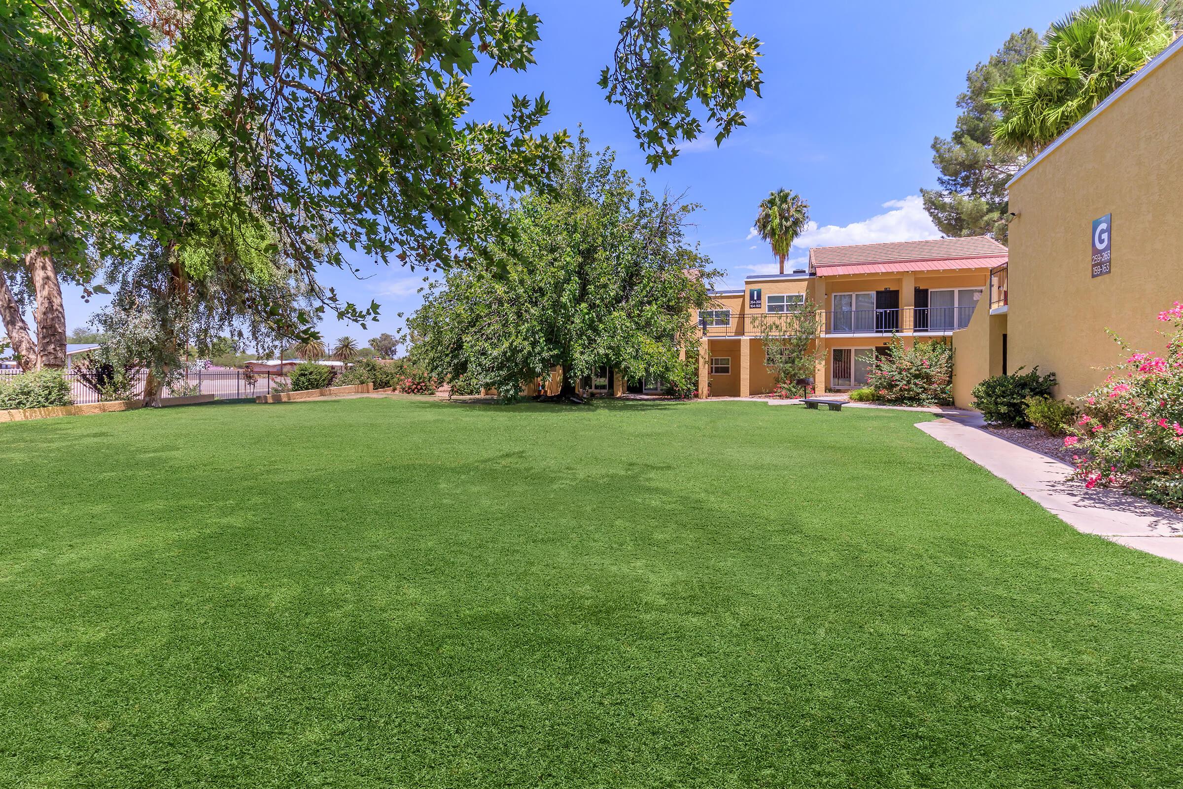 A vibrant green lawn surrounded by trees and flowering shrubs, with a building in the background featuring balconies. The sky is bright blue with a few clouds, creating a welcoming outdoor space.
