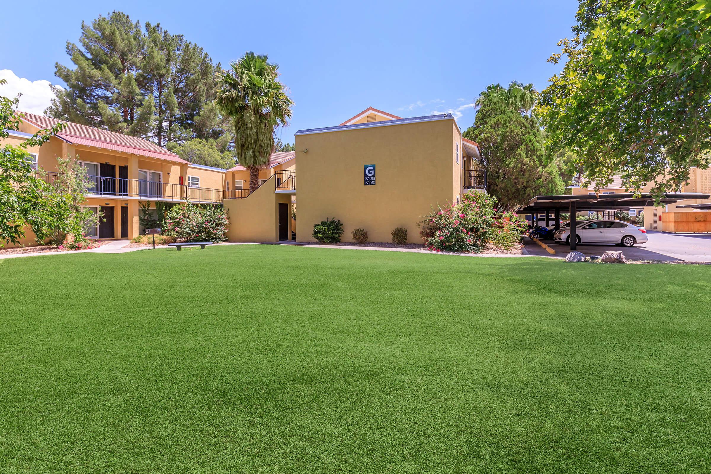 A well-maintained courtyard with artificial grass, surrounded by trees and shrubs. In the background, a yellow building with multiple entrances and balconies is visible. The area appears sunny and inviting, with a parking lot and cars in the background.