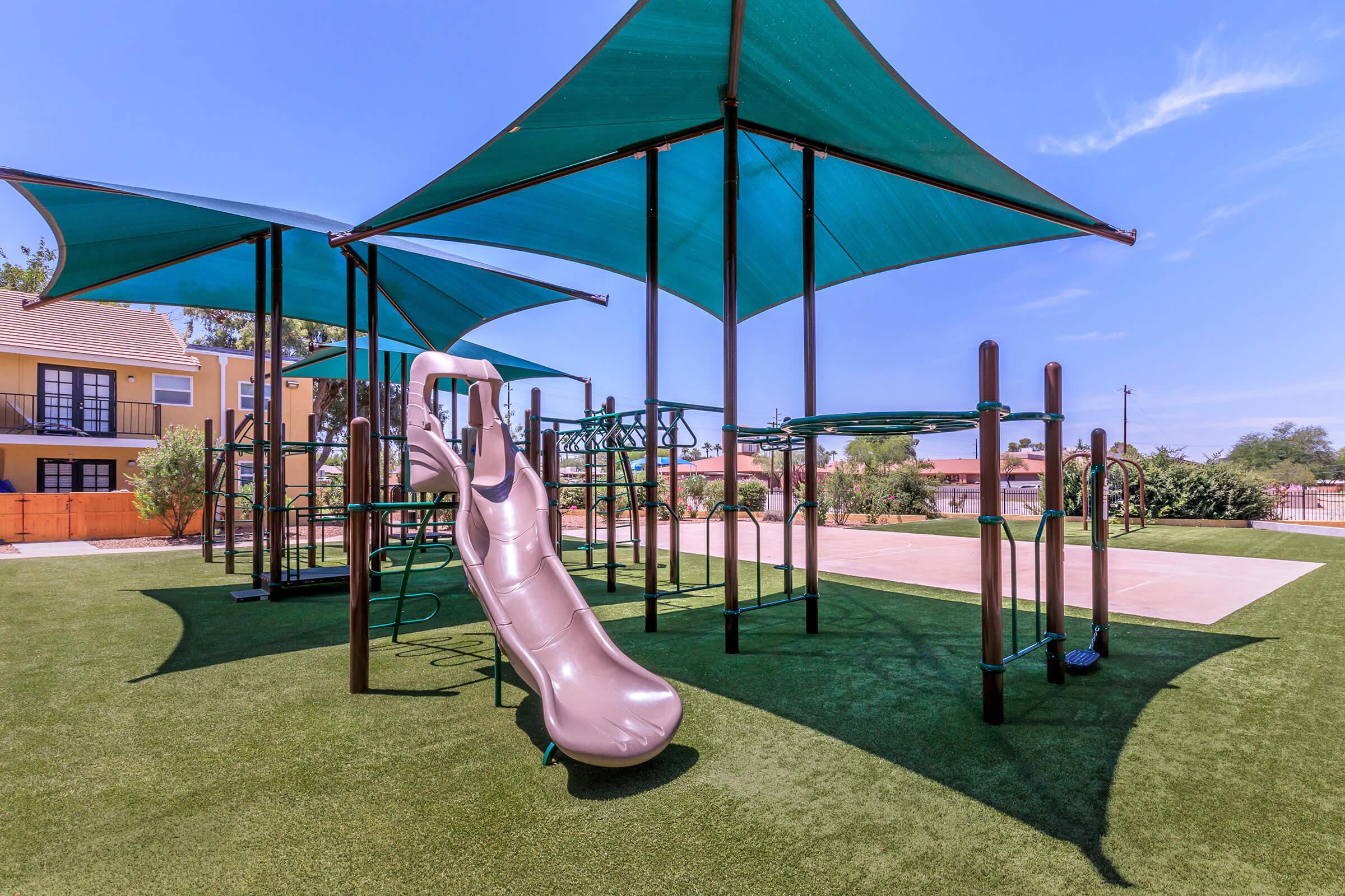 A playground featuring a brown slide, green climbing structures, and large aqua-colored shade sails. The area is covered with artificial turf, surrounded by greenery, with residential buildings visible in the background under a clear blue sky.
