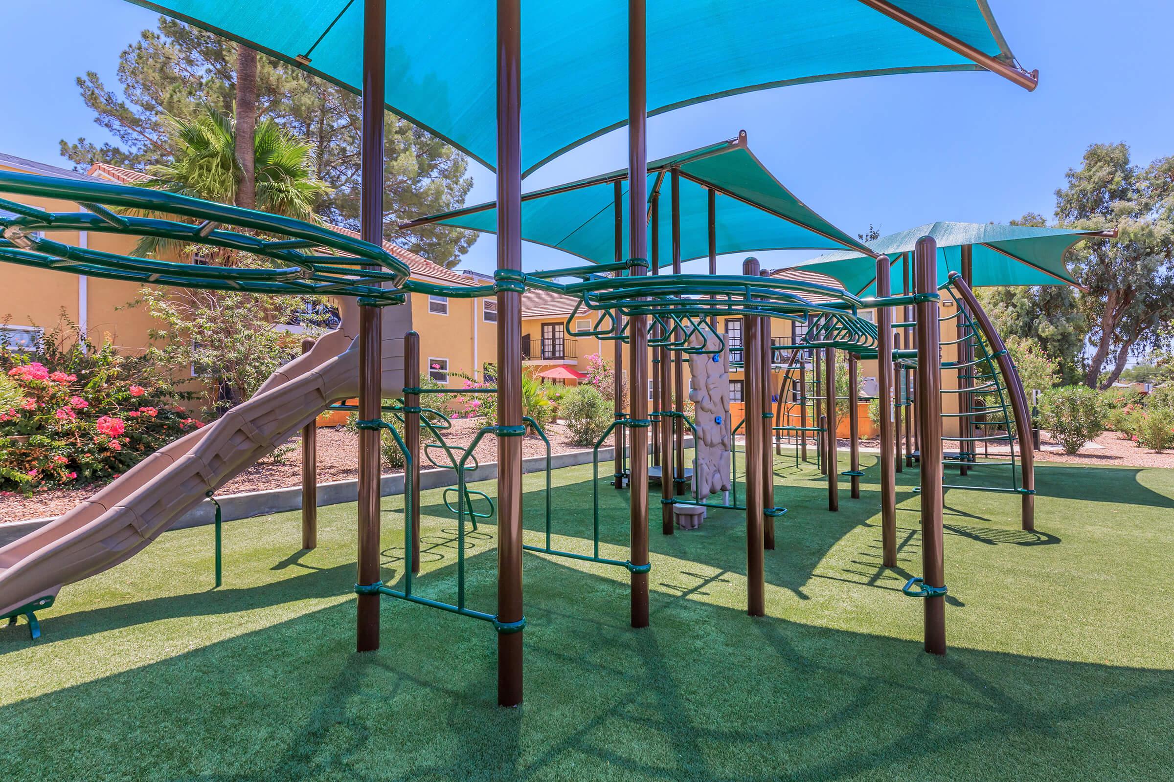 A colorful playground featuring climbing structures, a slide, and shaded areas under green canopies. The ground is covered with artificial grass, and there are blooming bushes in the background, creating a bright and inviting outdoor space for children to play.
