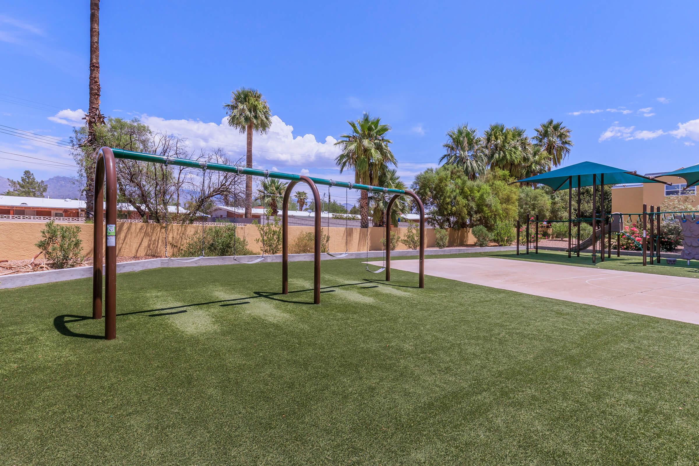 A playground featuring a set of swings and a slide, surrounded by lush green grass and palm trees. The sky is clear with a few clouds, creating a bright and inviting atmosphere for outdoor play.
