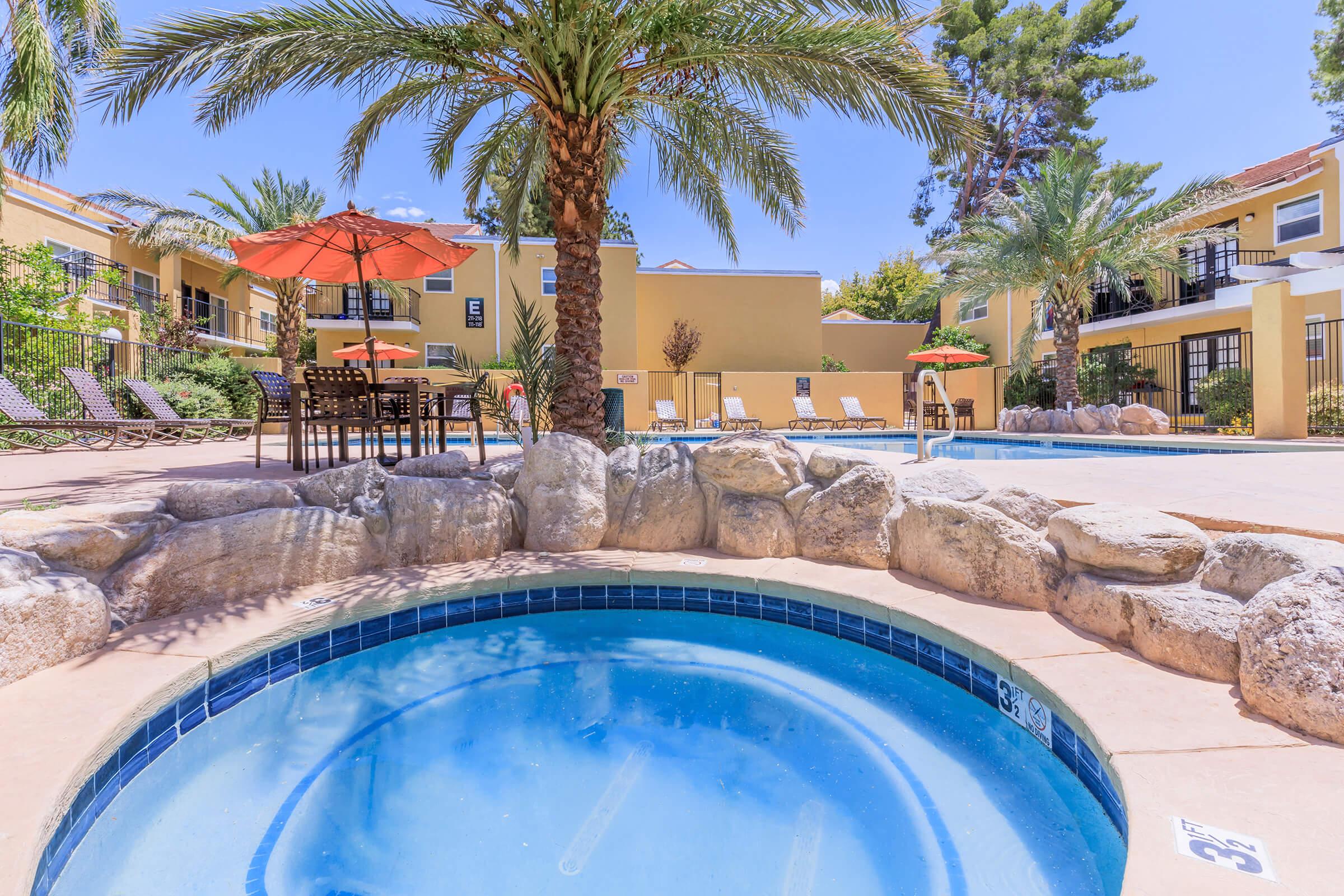 A swimming pool area featuring a hot tub in the foreground, surrounded by palm trees and rocks. Lounge chairs and umbrellas are arranged around the pool, with a sunny blue sky overhead. The building in the background adds a welcoming ambiance to the outdoor space.