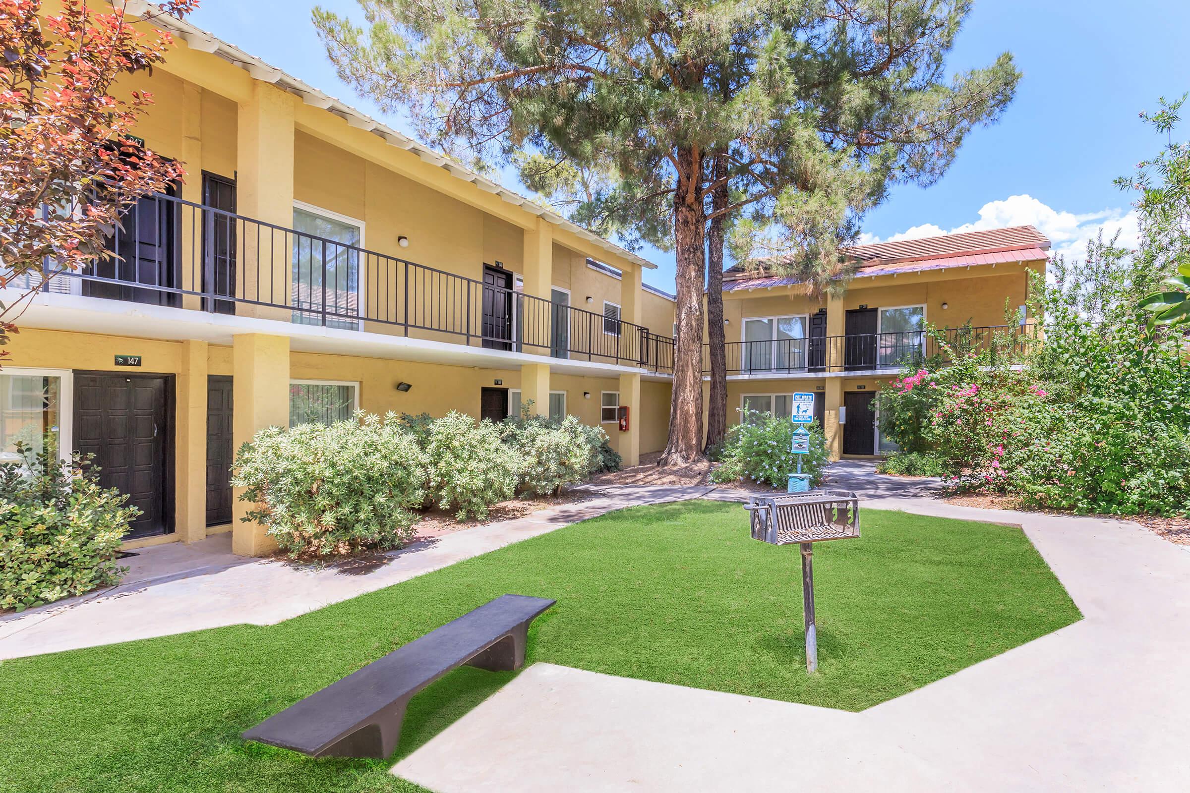 A landscaped courtyard featuring two apartment buildings with balconies, surrounded by greenery and trees. There is a grassy area in the center with a bench and a BBQ grill, creating a relaxed outdoor space. The sky is clear with a few clouds, suggesting a sunny day.
