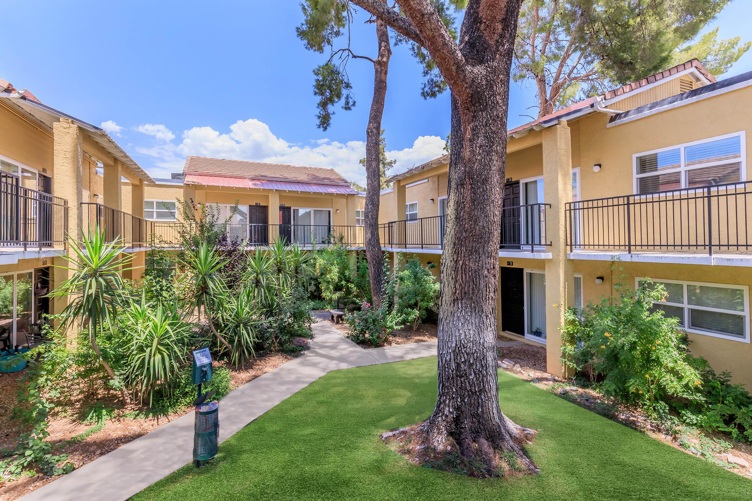 A well-maintained courtyard surrounded by two-story apartment buildings. Lush green plants and trees provide shade, with a curved pathway leading through the garden area. The sky is bright blue with a few clouds, creating a serene and inviting atmosphere.