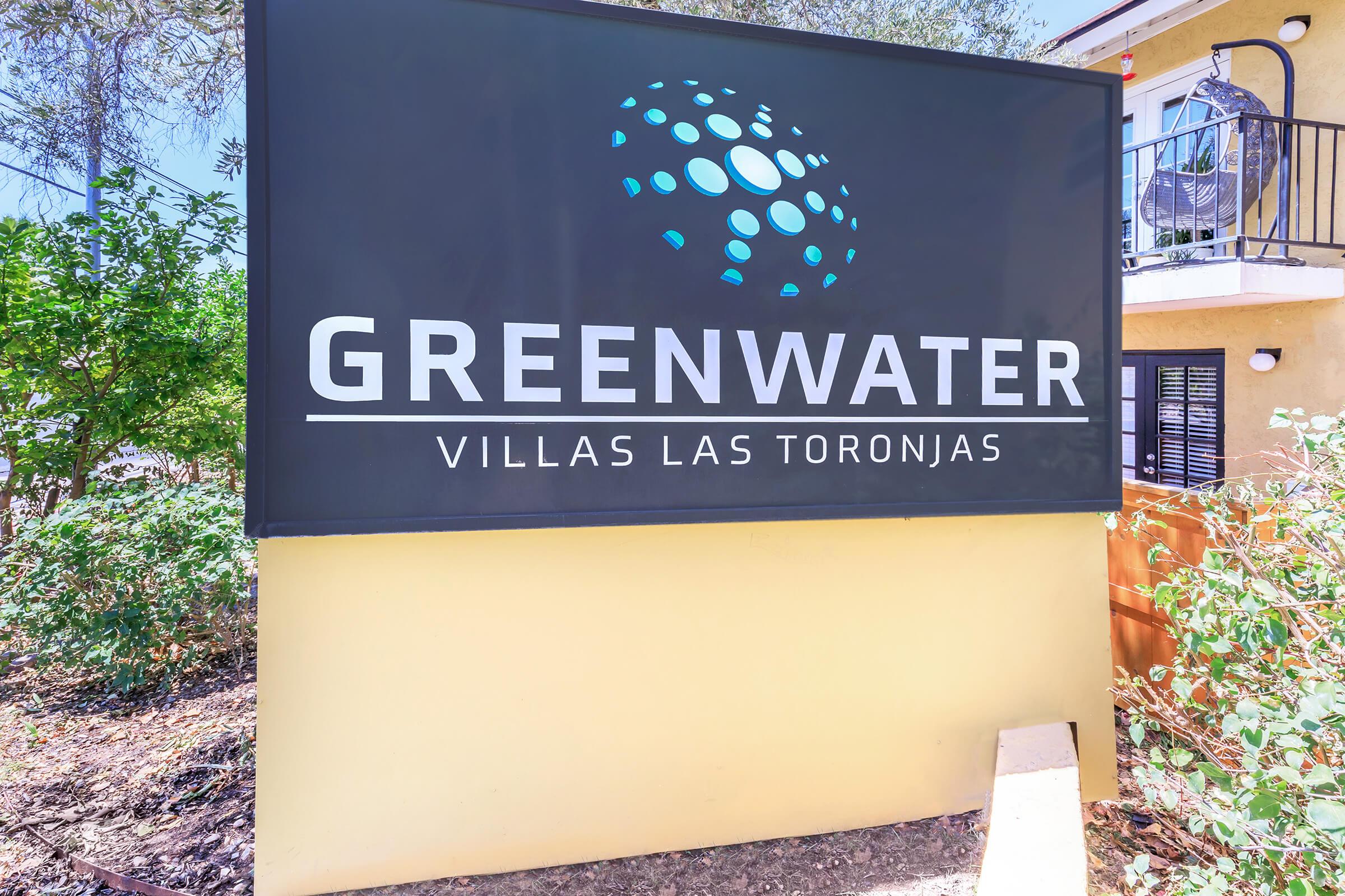 Sign for Greenwater Villas Las Toronjas, featuring a modern design with a blue and white logo on a black background. The sign is surrounded by greenery and is positioned near a building with balconies.