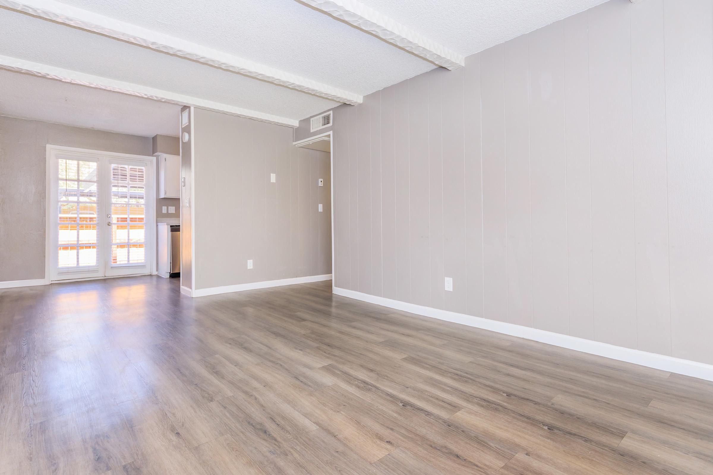 Spacious interior of an empty room with light-colored walls and wooden flooring. Features include exposed ceiling beams and a sliding glass door leading to a patio or exterior space. The area is well-lit with natural light coming in through the door.