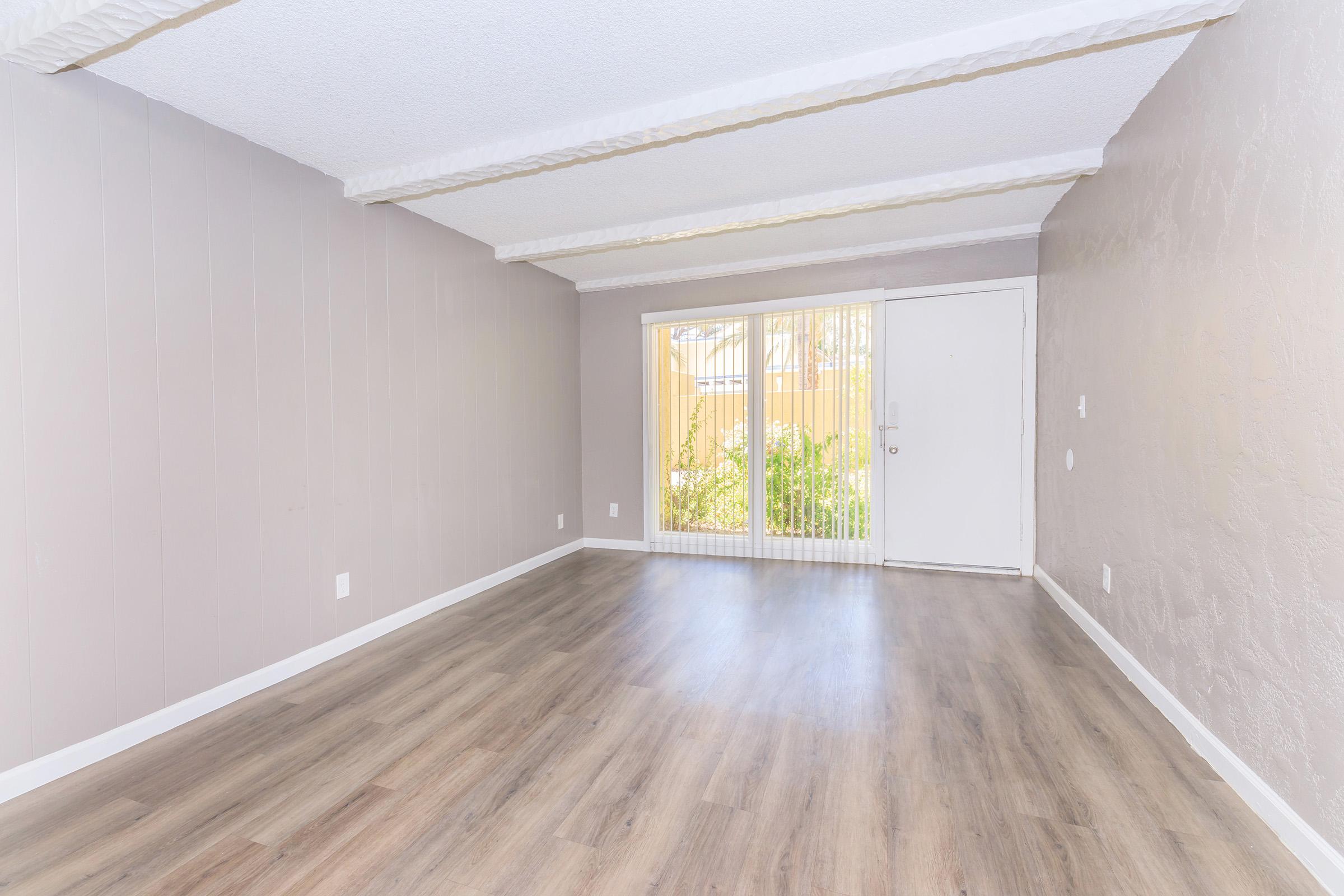 An empty room with light-colored walls and a textured ceiling. The floor has wood-like flooring. There is a sliding glass door at the back, leading to a patio area with visible greenery. The room is well-lit, with natural light coming through the door.
