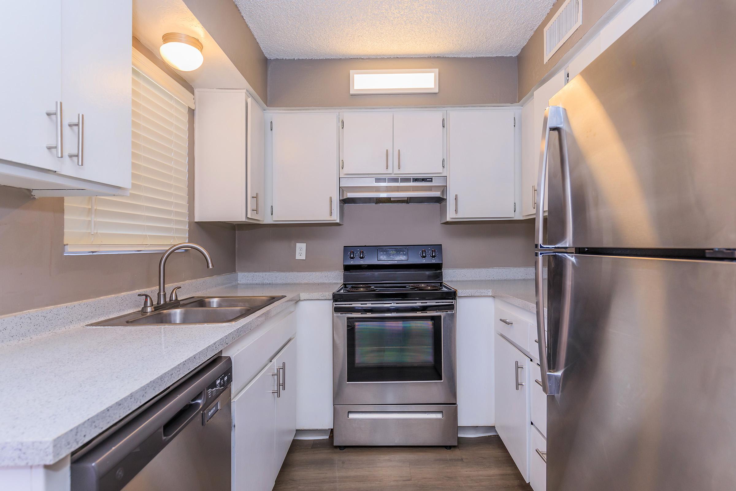 Modern kitchen featuring white cabinets, stainless steel appliances including an oven, stove, and refrigerator. The countertop is grey with a subtle texture, and there's a double sink beside a window. Soft lighting illuminates the space, enhancing the clean and contemporary design.