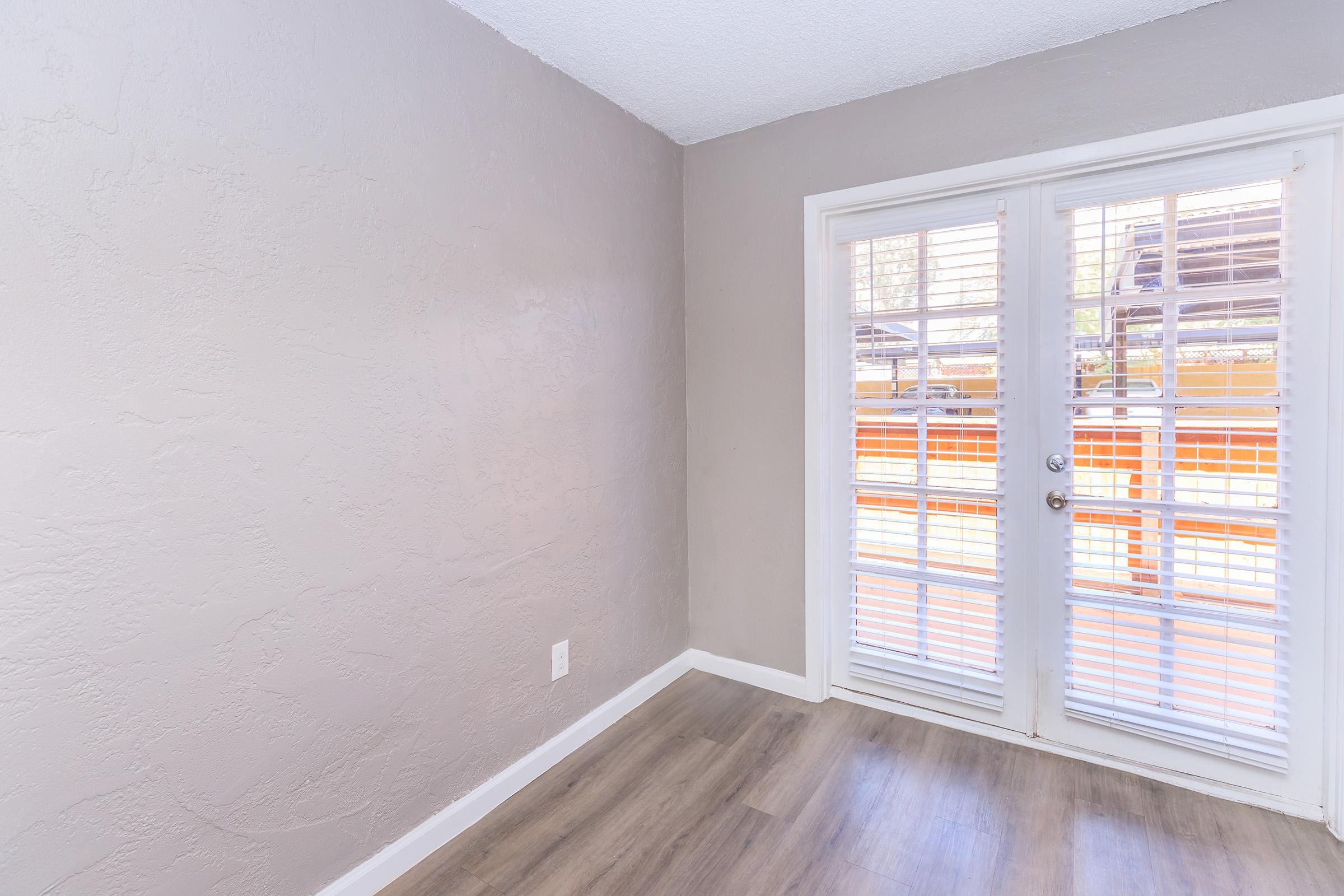 Empty room with light gray walls and a smooth wooden floor. There are two glass doors with white blinds that open to an outdoor area, providing natural light. The atmosphere is bright and neutral, suitable for various uses.