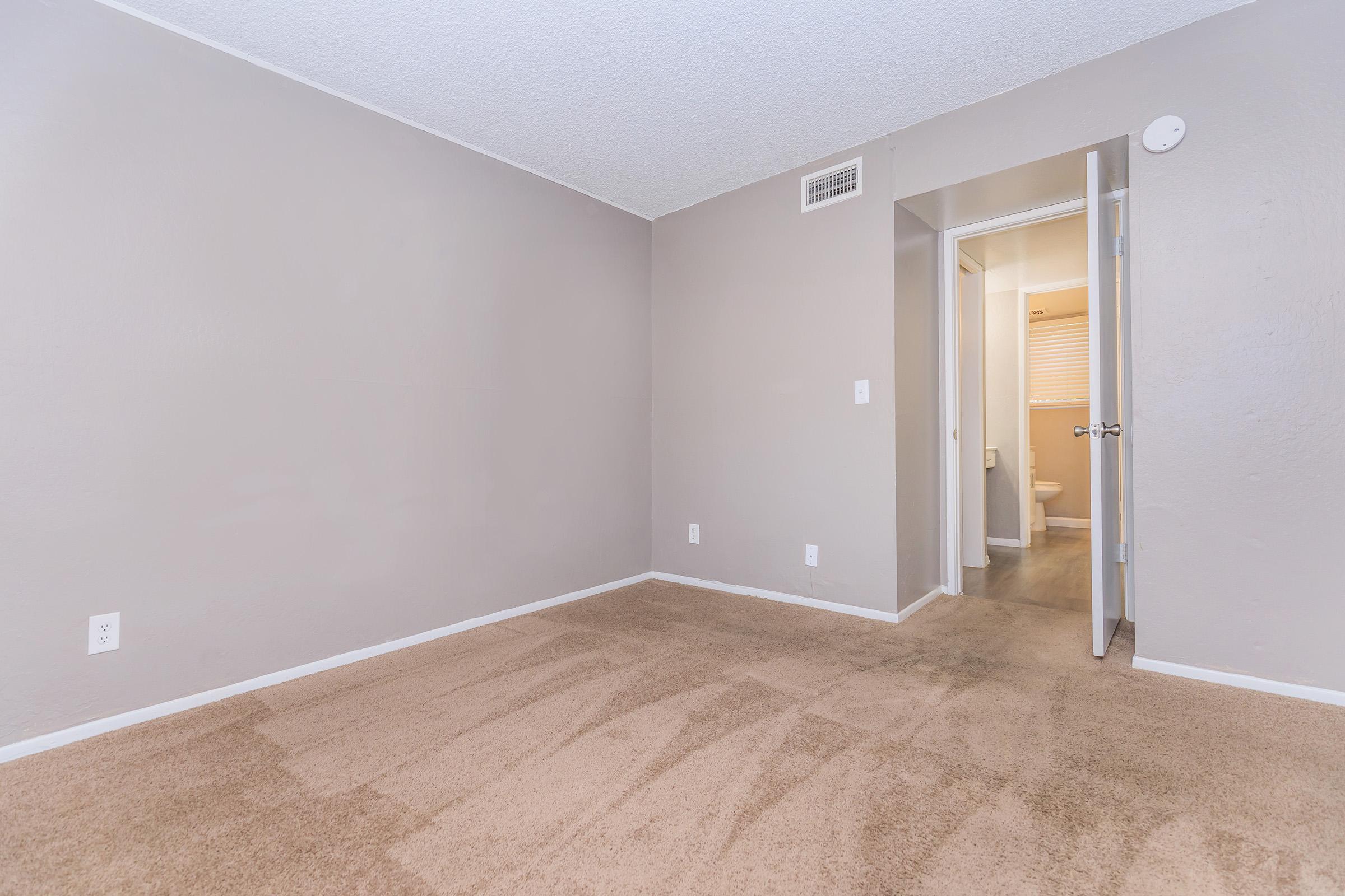 Empty room with beige carpet and light gray walls. A door is open in the background leading to a bathroom area with a light source visible. The space is uncluttered, showcasing the room's layout and dimensions.