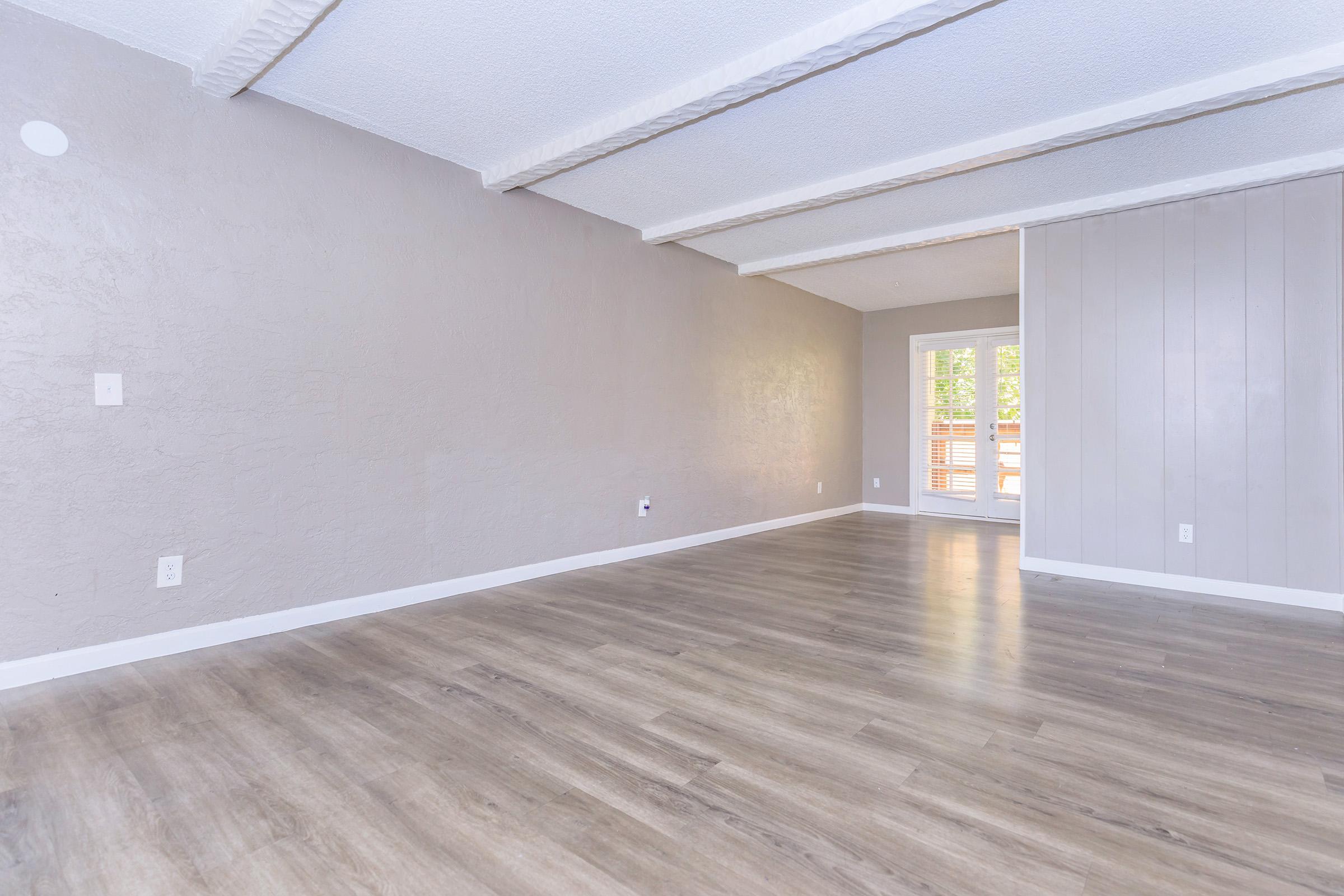 An empty room with gray walls and wooden flooring, featuring exposed ceiling beams. A window with vertical blinds allows natural light to enter from the right side of the image. The space has a minimalist design, with no furniture or decorations present.