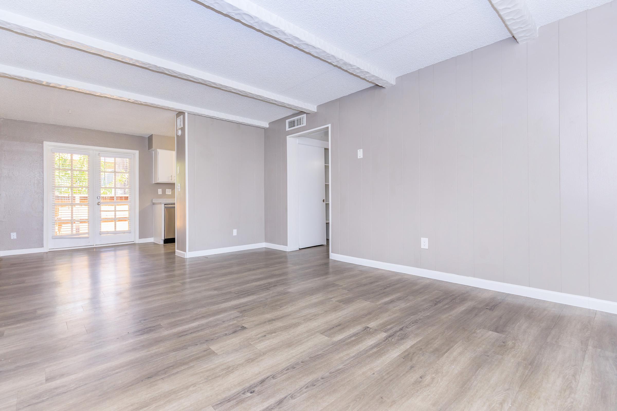 A spacious, empty room with light-colored walls and wooden flooring. The room features exposed beams on the ceiling and an open layout leading to a kitchen area with a doorway visible. Sunlight streams in from a set of sliding glass doors that lead to a patio or balcony outside.