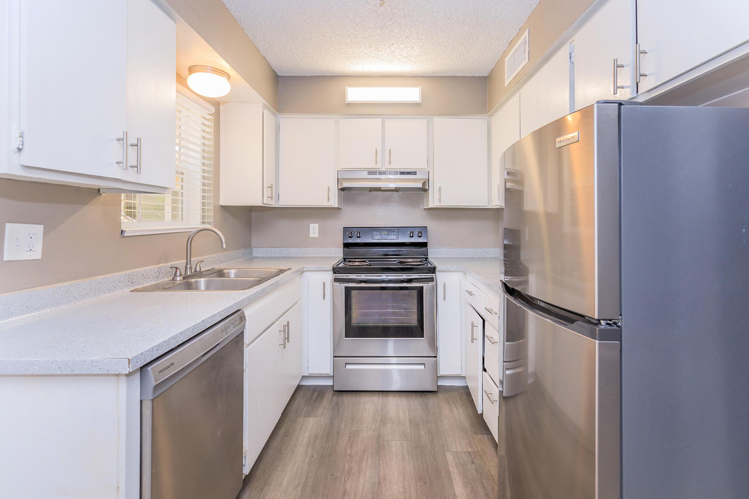 Modern kitchen featuring stainless steel appliances, including a refrigerator, oven, and dishwasher. Counters are light-colored with a smooth finish, and cabinets are white. The room is well-lit with natural light from a window and a ceiling light. The floor has a wood-like appearance, enhancing the contemporary look.