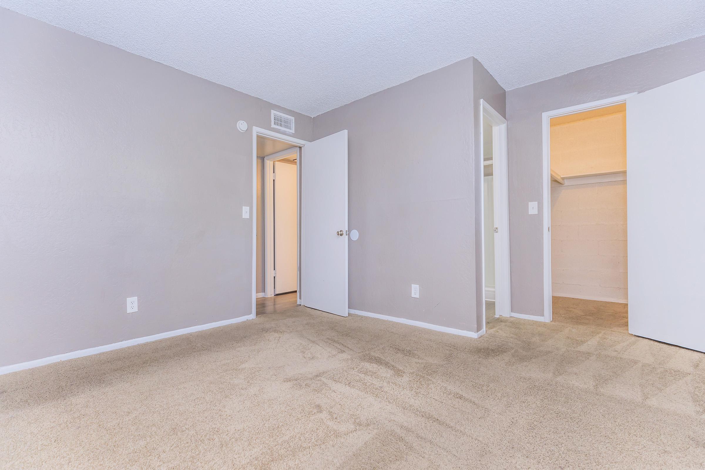 A spacious, empty room with beige carpet and light gray walls. Two closed doors are visible, leading to adjacent areas, and one door is slightly ajar. The room has a clean and simple design, providing a blank canvas for decorating. Natural light filters in through the open door.