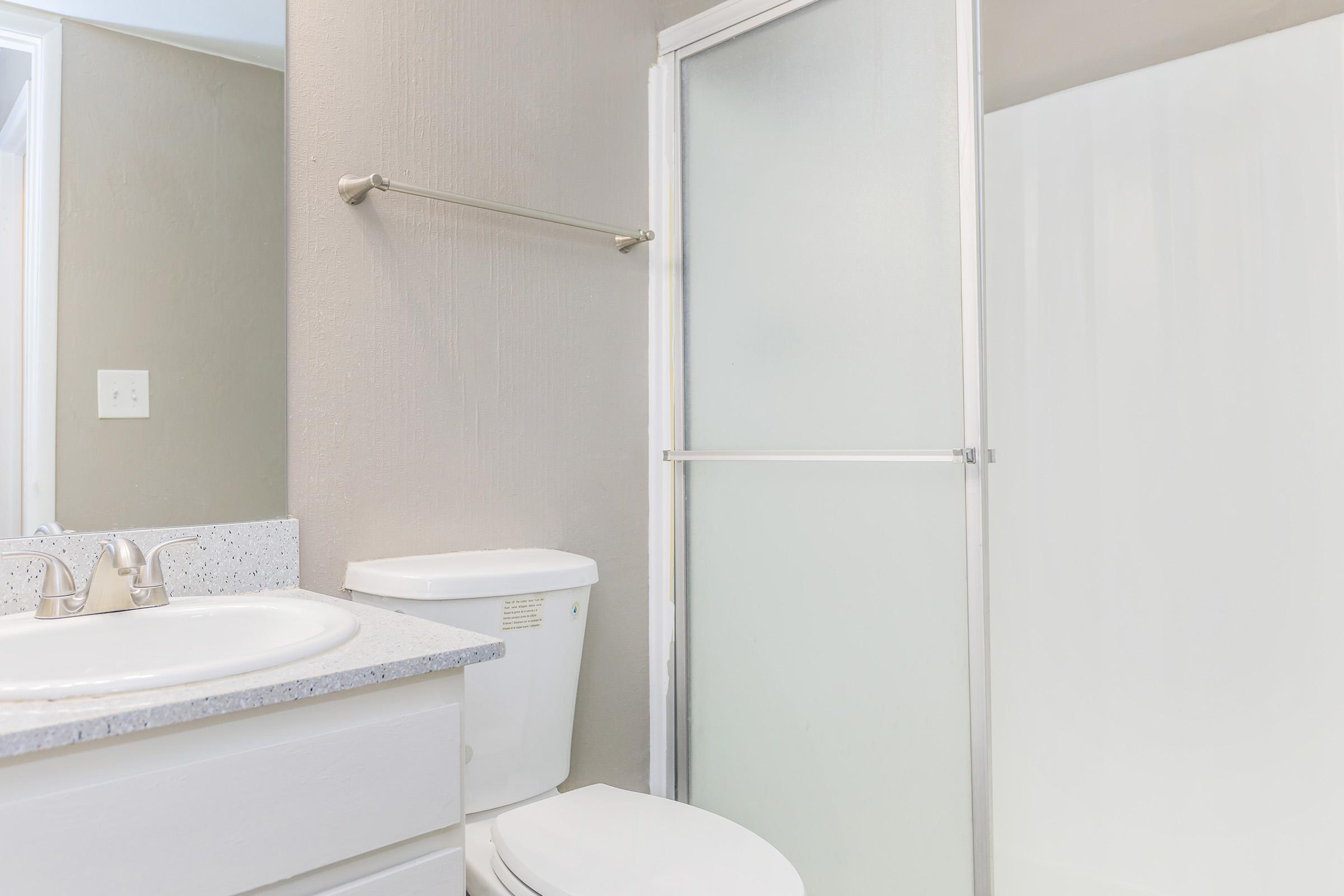 A clean bathroom featuring a white toilet, a sink with a countertop, and a glass shower door. The walls are a neutral shade, and there's a towel bar mounted above the sink. Natural light enters from an adjoining room.