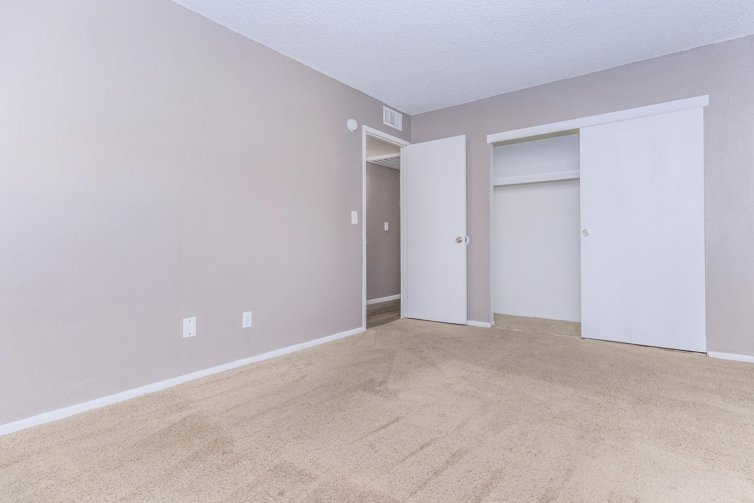 Empty room with light gray walls and beige carpet. There are two white sliding closet doors on the right and a door leading to another space on the left. The room appears spacious and well-lit, with a simple and clean design.