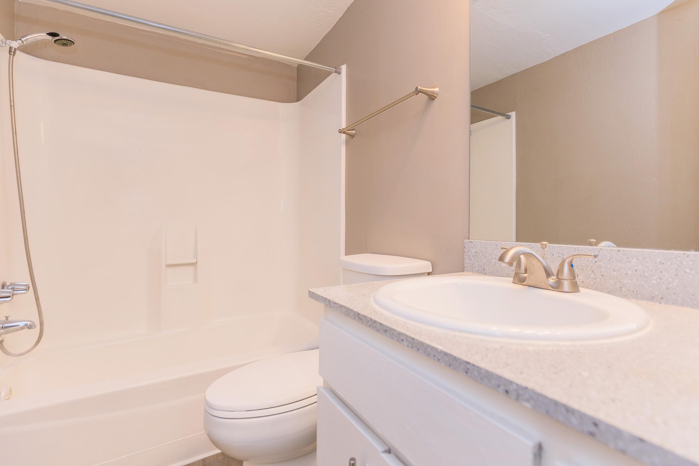 A clean and modern bathroom featuring a white bathtub and shower combination, a light-colored countertop with a sink, and a simple mirror. The walls are painted in a soft beige, enhancing the spacious feel of the area. The overall design is minimalistic and well-lit.