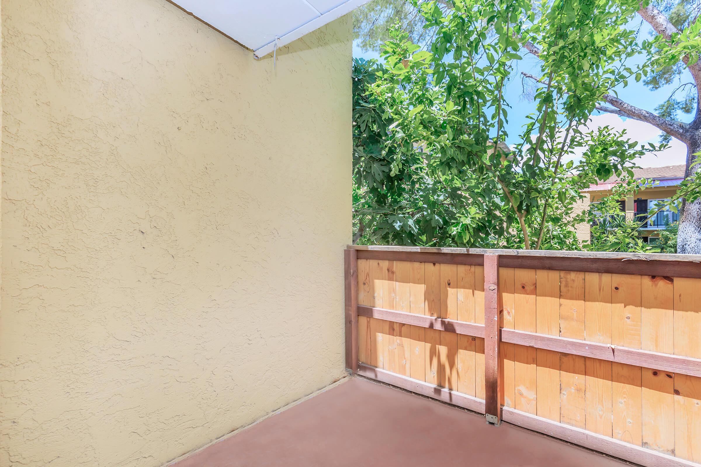 A small balcony or patio with a wooden railing, surrounded by greenery. The wall is painted a light yellow, and there is a clear blue sky visible beyond the trees. The space appears bright and inviting, suitable for relaxation or enjoying the outdoors.