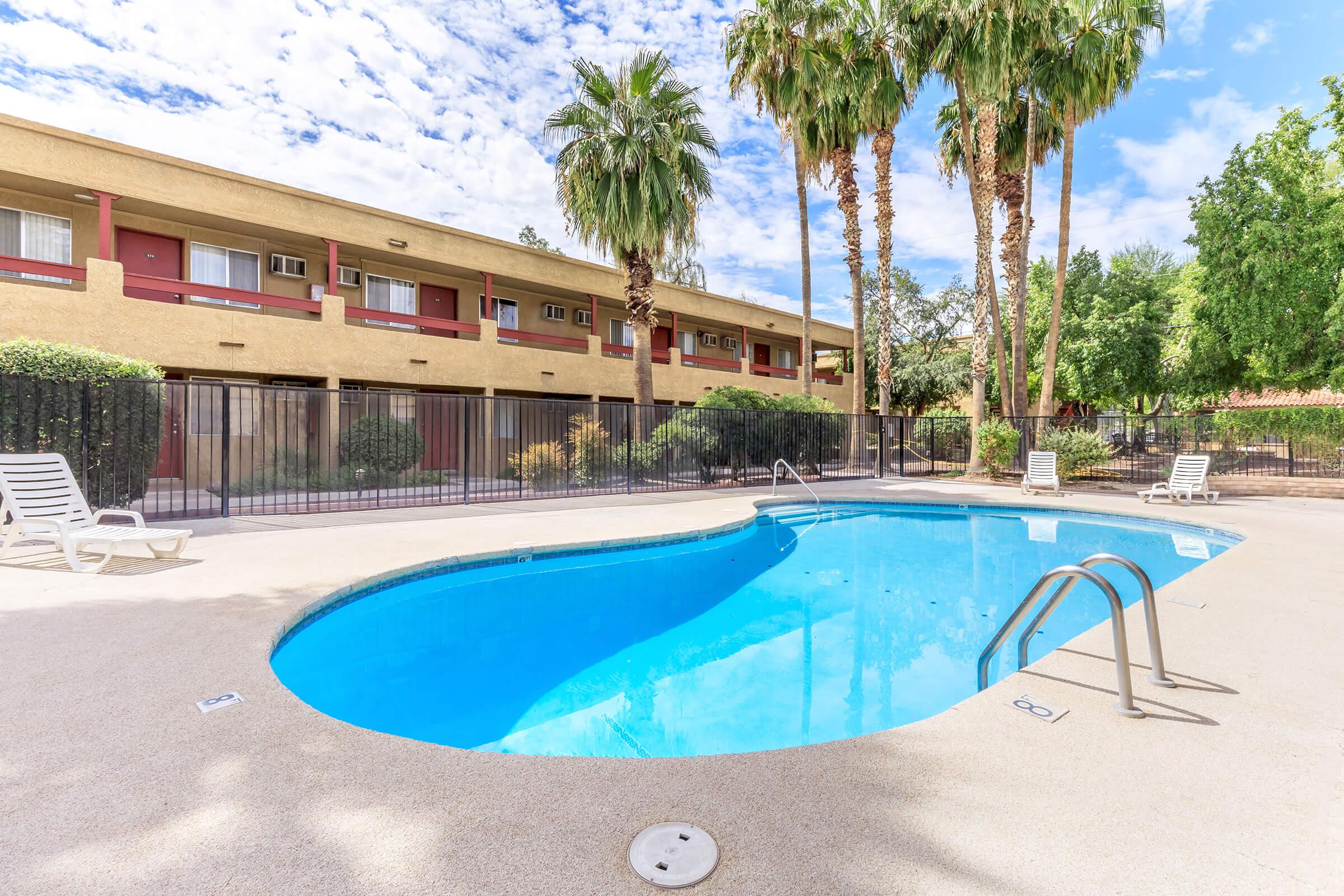 A clear blue swimming pool surrounded by palm trees and neatly landscaped greenery, with lounge chairs positioned nearby. In the background, a two-story building with red accents and multiple windows is visible under a partly cloudy sky.