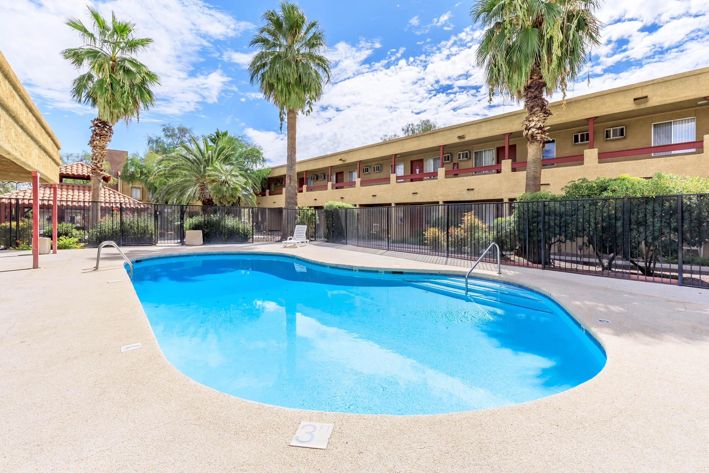 A glistening blue swimming pool surrounded by palm trees and a fence. In the background, a beige two-story building with balconies is visible under a partly cloudy sky. The pool area features a clean deck and is well-maintained, creating a relaxing atmosphere.
