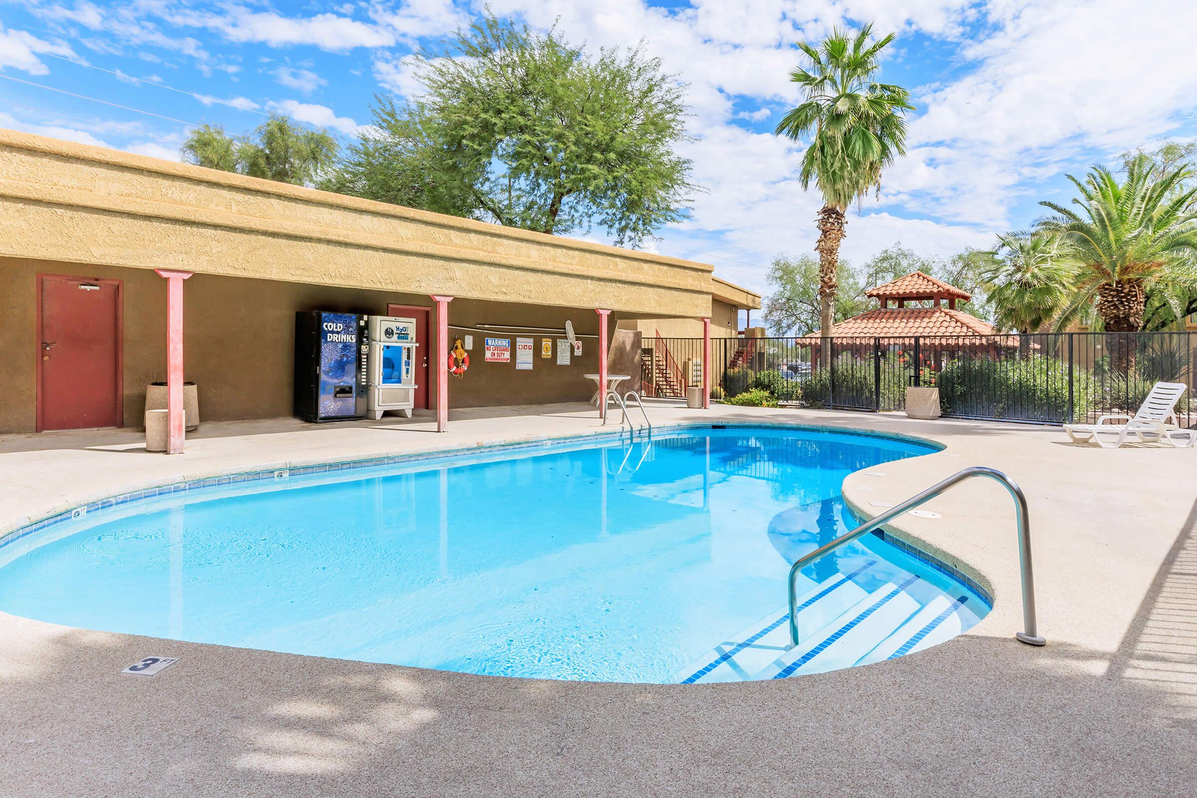 A serene outdoor swimming pool with a clear blue surface, surrounded by palm trees and a gated area. Lounge chairs are positioned nearby, and a vending machine is visible. The sky above is partly cloudy, adding to the tranquil atmosphere.