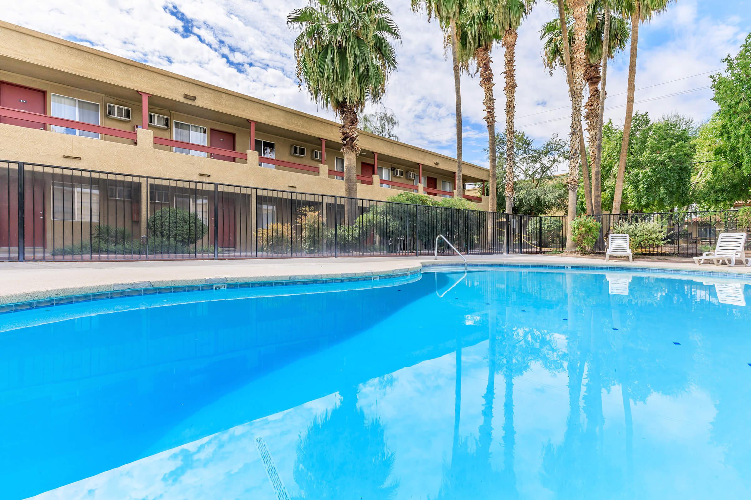 View of a sparkling blue swimming pool surrounded by a black fence, with palm trees in the background. An apartment building with a cream-colored exterior and red accents is visible in the background, reflecting in the water. The sky is partly cloudy, adding to the serene atmosphere.