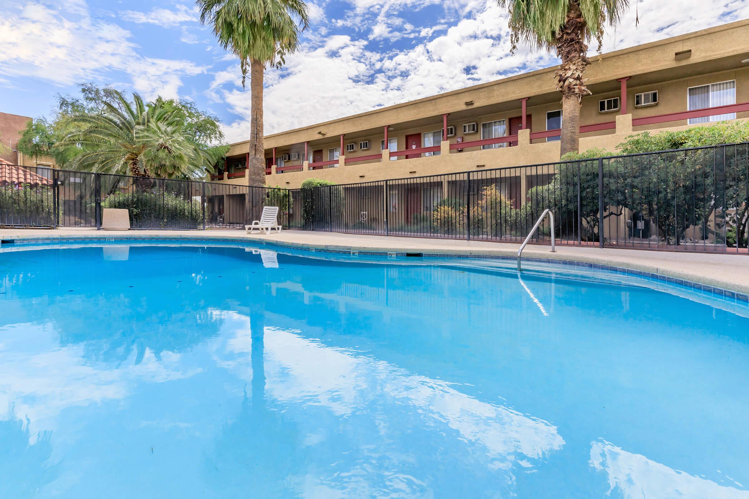A tranquil swimming pool surrounded by palm trees, with a two-story building in the background. The sky is partly cloudy, reflecting blue tones on the water's surface. A lounge chair is positioned nearby, creating a relaxed atmosphere for leisure and enjoyment.