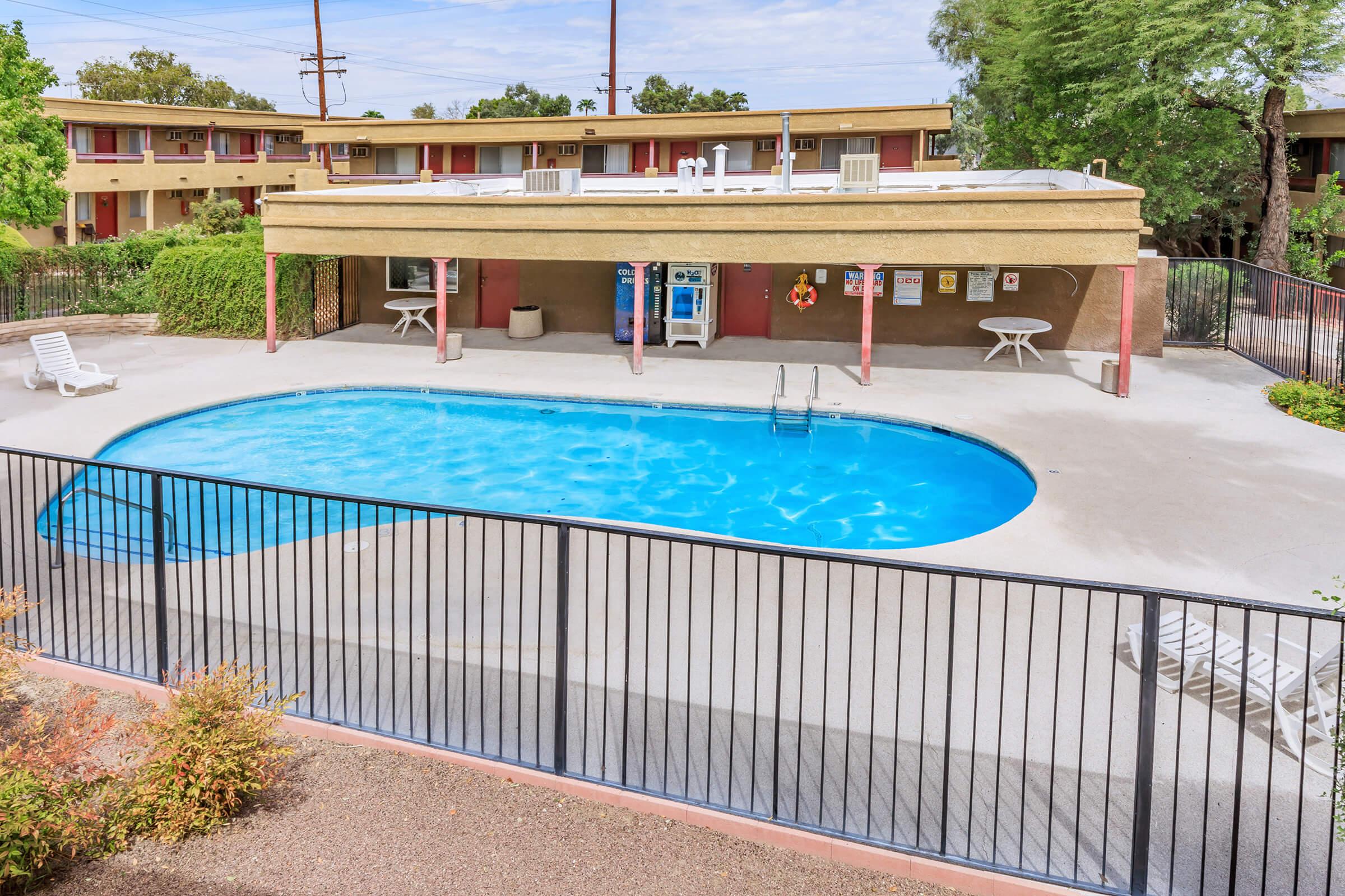 A clean, inviting swimming pool surrounded by a fence. The pool area features several lounge chairs and a shaded seating area with a table. In the background, there are two-story buildings with balconies. Lush greenery surrounds the pool, creating a relaxing atmosphere.