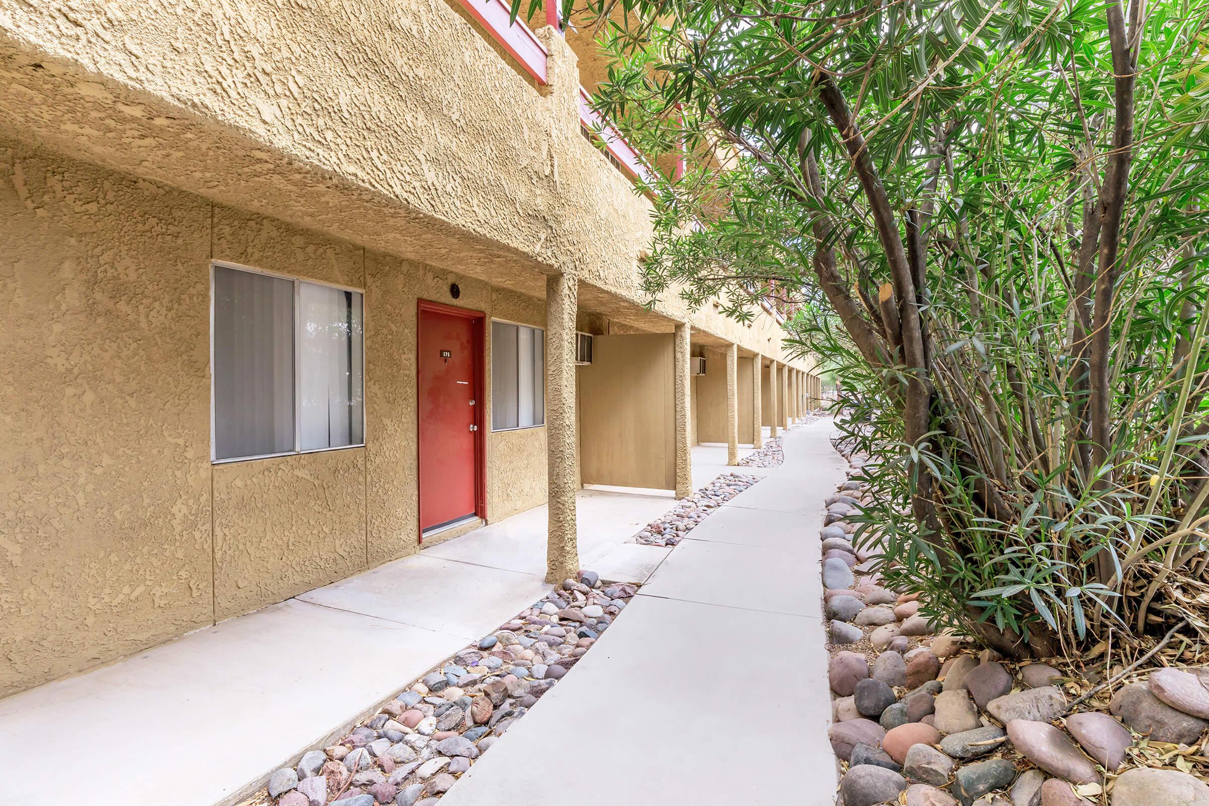 A pathway runs alongside a series of beige-colored buildings with red doors. Lush greenery, including bamboo-like plants, lines one side of the walkway. The scene depicts a well-maintained outdoor area of a residential or commercial building complex.