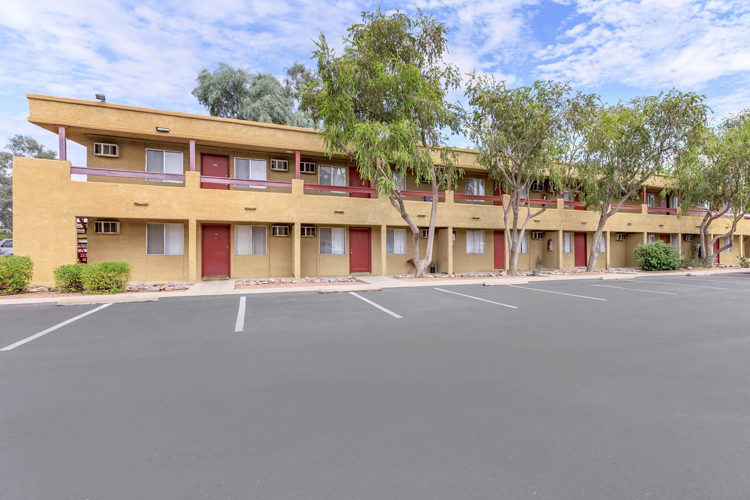 A view of a two-story motel with tan walls and red doors, surrounded by trees. The parking lot in front is empty, and the sky is partly cloudy. The building features multiple rooms with a balcony style, creating a simple and functional design.