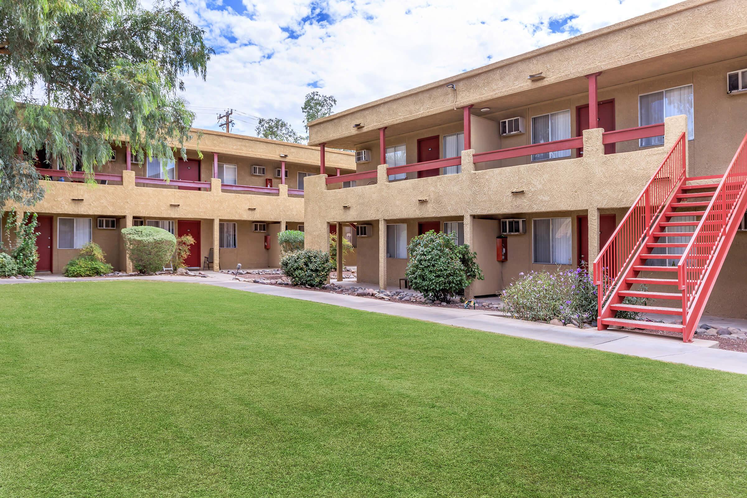A view of a tidy apartment complex featuring two-story buildings with balconies. The buildings have a beige exterior and red accents, surrounded by well-maintained green lawns and shrubs. A staircase leads to the second floor, and the sky is partly cloudy.